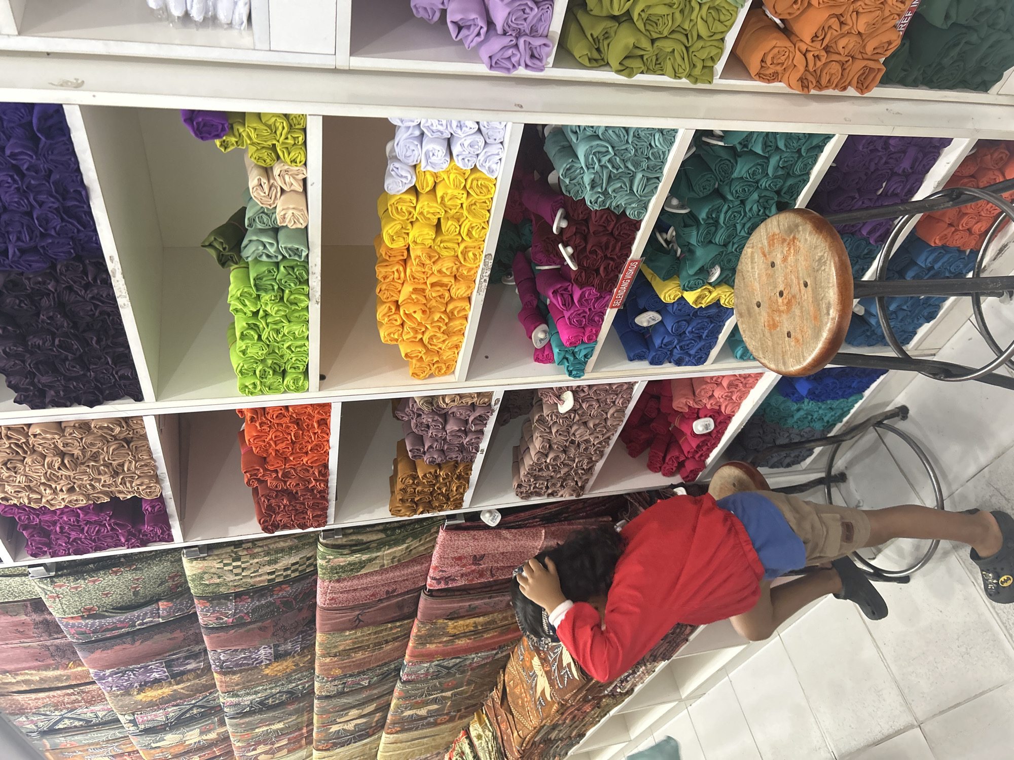 A young child, wearing a red shirt and beige shorts with a blue undershirt, is seen from behind, lying on stacks of colorful textiles. The child is resting their head on their hands, appearing relaxed or perhaps tired, amidst a retail setting. The shelves behind the child are filled with tightly rolled fabrics in a vibrant spectrum of colors, including purple, black, lime green, yellow, orange, teal, maroon, and blue. Below these shelves, more fabric is displayed, featuring intricate patterns and designs. A round, wooden stool with visible holes sits on a metal chair frame, suggesting a shop or market stall. The lighting is bright and even, indicating an indoor environment, possibly a fabric store in Ubud District, Indonesia. The overall scene suggests a moment of rest or play in a busy commercial space.