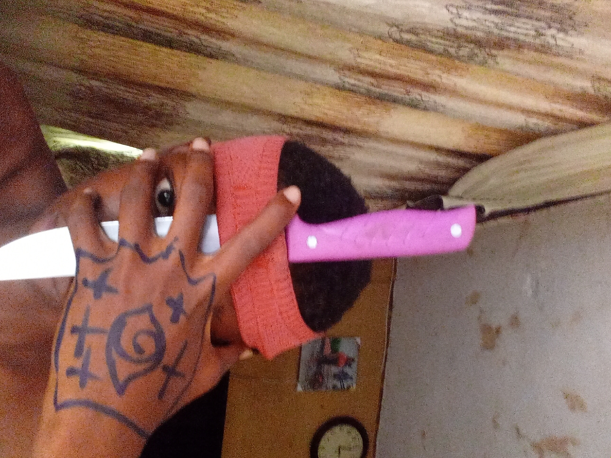 An individual with dark skin holds a pink-handled knife. The hand displays a drawing with cross-shaped symbols. A red fabric material covers part of the object held. The scene includes a wall with a picture and a clock. The location is Onitsha, Nigeria.