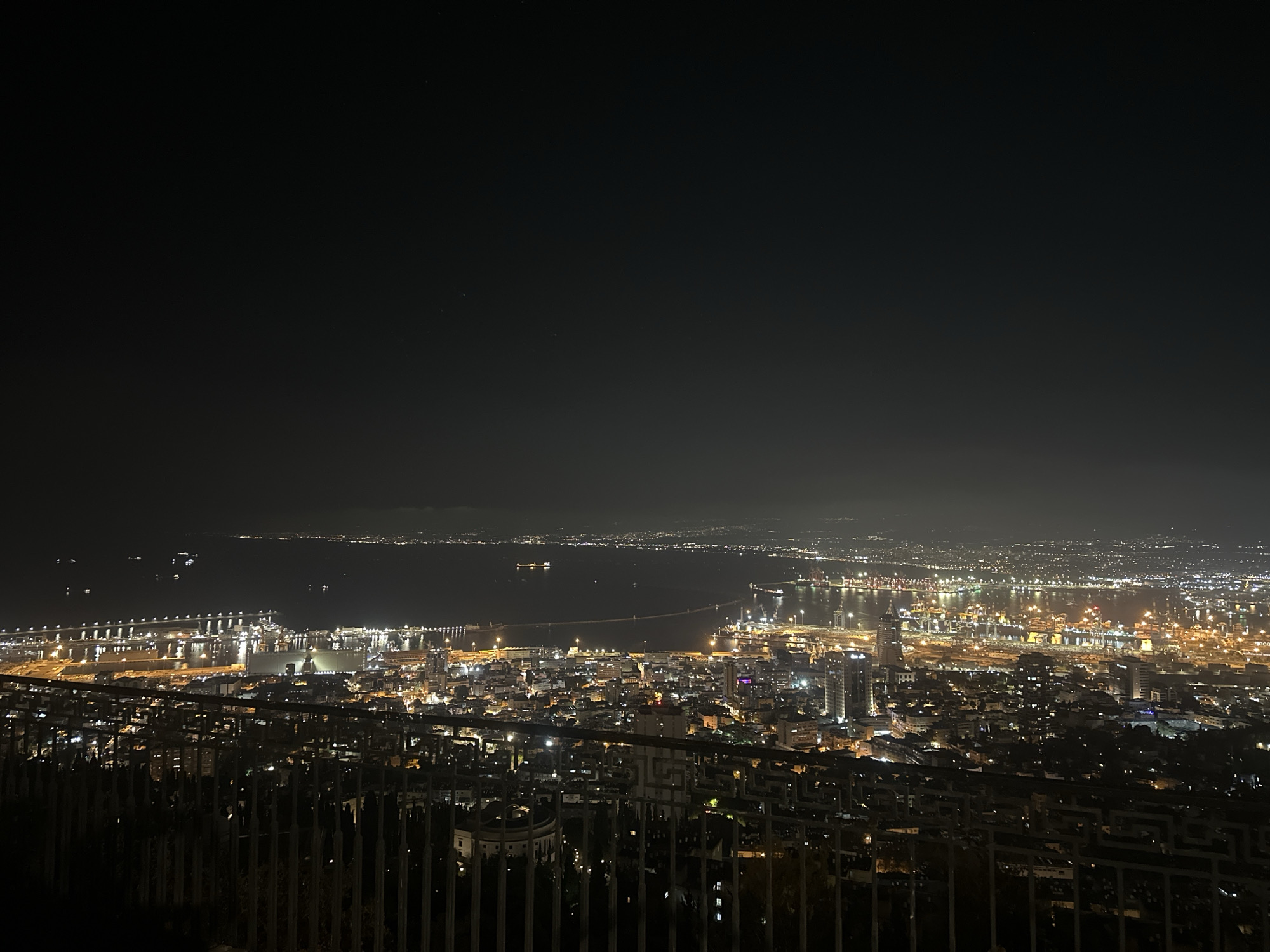 An elevated nighttime panoramic view shows the city of Haifa, Israel, extending towards the Mediterranean Sea. In the foreground, a dark metal railing is visible, with sparse vegetation and architectural elements partially illuminated below. The city lights of Haifa create a bright, sprawling tapestry below. Dominating the mid-ground is the extensive port complex, characterized by numerous brightly lit structures, cranes, and docked ships. The harbor is filled with various vessels, some appearing to be cargo ships and others smaller boats, their lights reflecting on the water. Further out, the dark expanse of the sea stretches towards a distant, faintly lit coastline, possibly indicating other settlements. The sky above is dark, with a subtle gradient of lighter tones near the horizon, suggesting ambient light pollution from the city. The overall scene captures the active maritime and urban life of Haifa at night.
