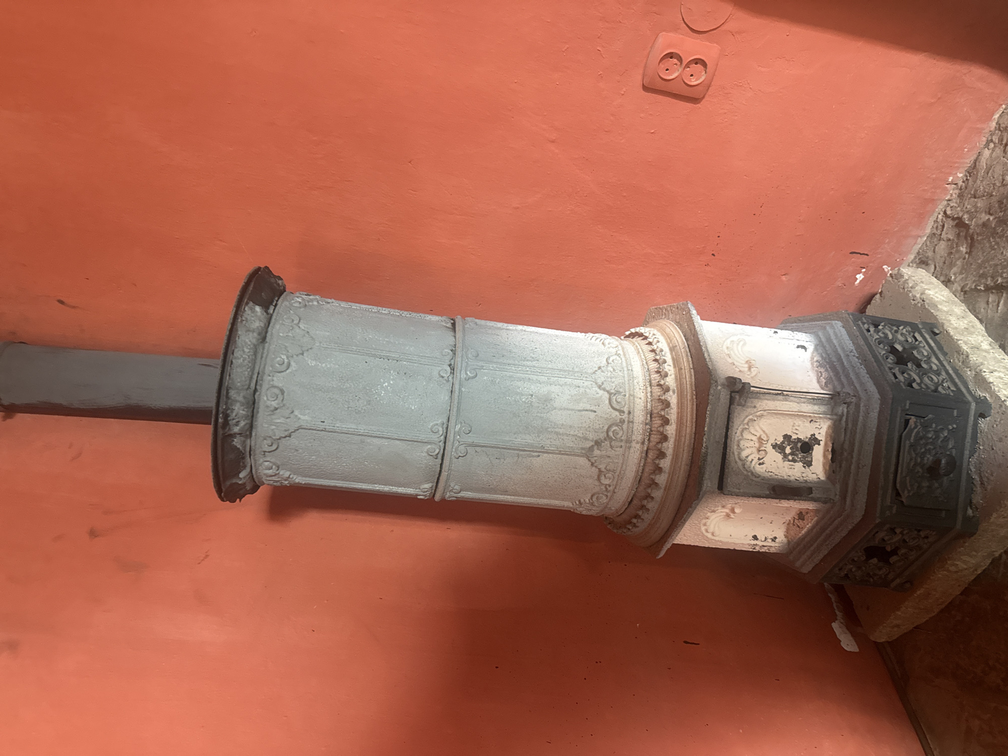 A horizontal shot depicts a vintage or antique white cylindrical stove with ornate detailing, resting against a salmon-colored wall. The stove extends towards the left side of the frame, with a gray pipe protruding from its left end. The right end of the stove rests on a beige, possibly stone, base. A European electrical outlet is visible on the wall in the upper right corner. The image may have been taken in Chynadiiovo, Ukraine.
