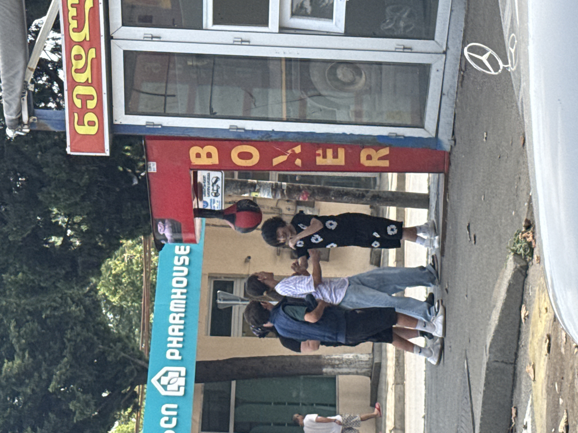 In Tbilisi, Georgia, on a sunny day, a group of four individuals stands on a paved sidewalk near a building. A male and a female, appearing to be teenagers or young adults, face each other, with the female in a black dress with a floral pattern and the male in a blue vest over a dark shirt and dark shorts. They are positioned near a red and blue structure with the word "BOXER" vertically displayed in yellow letters. A black and red punching bag hangs from the structure. To their left, two other individuals are visible. One, wearing a blue vest and dark shorts, stands partially obscured, and another, in a white and purple striped shirt and light blue jeans, stands behind the male in the vest. The background features a building with a turquoise sign that reads "PHARMHOUSE" and trees. The scene is captured from a low angle, with a portion of a white vehicle in the foreground and a drawing of a Mercedes-Benz logo on the pavement.