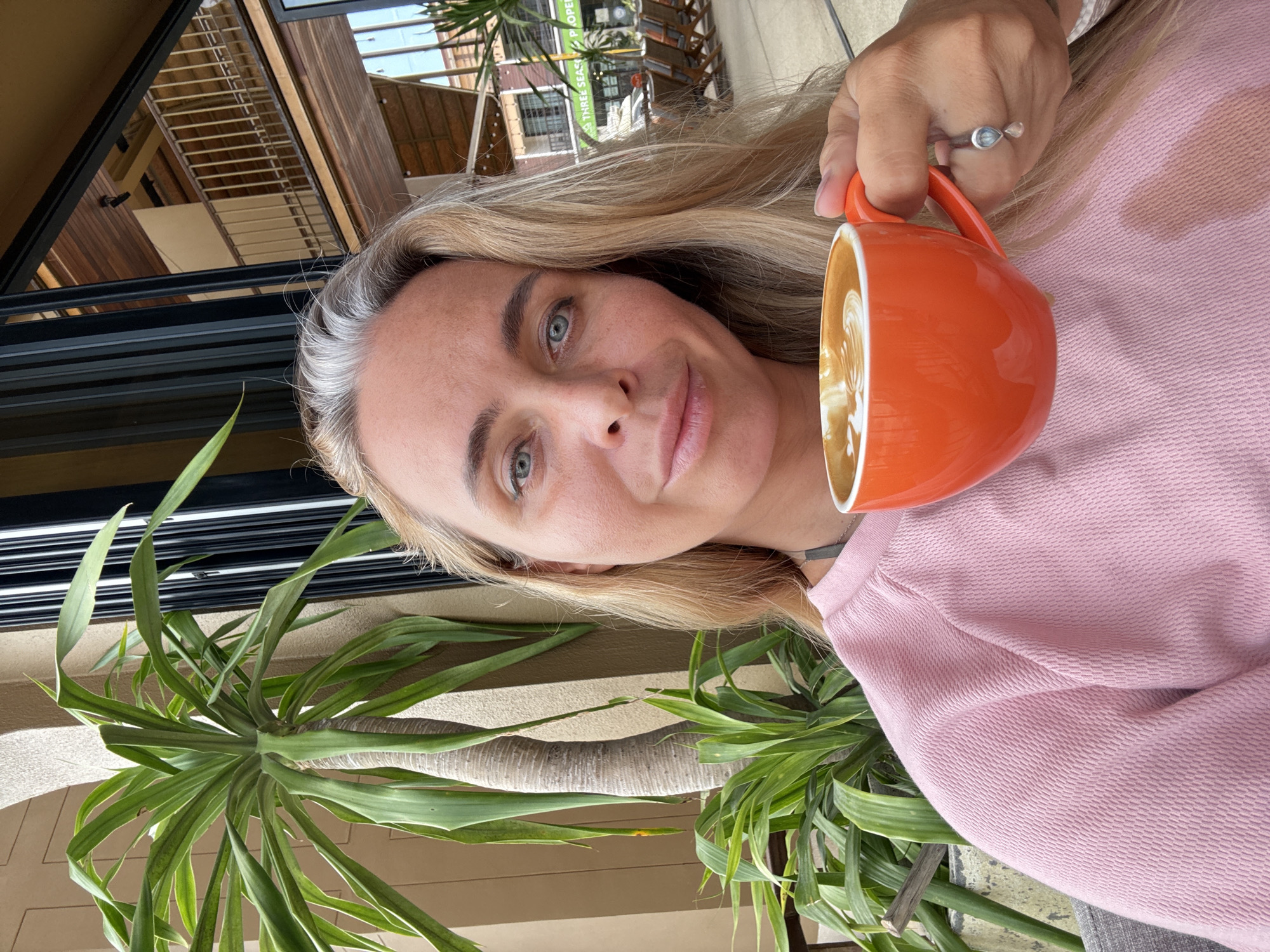 A woman with blond hair and blue eyes is taking a selfie while holding an orange mug of coffee. She is wearing a pink sweater. She is sitting outside, with a potted plant and a building visible in the background. The building appears to be a cafe or restaurant, with outdoor seating. The city of Ko Samui District, Thailand is mentioned as the location.