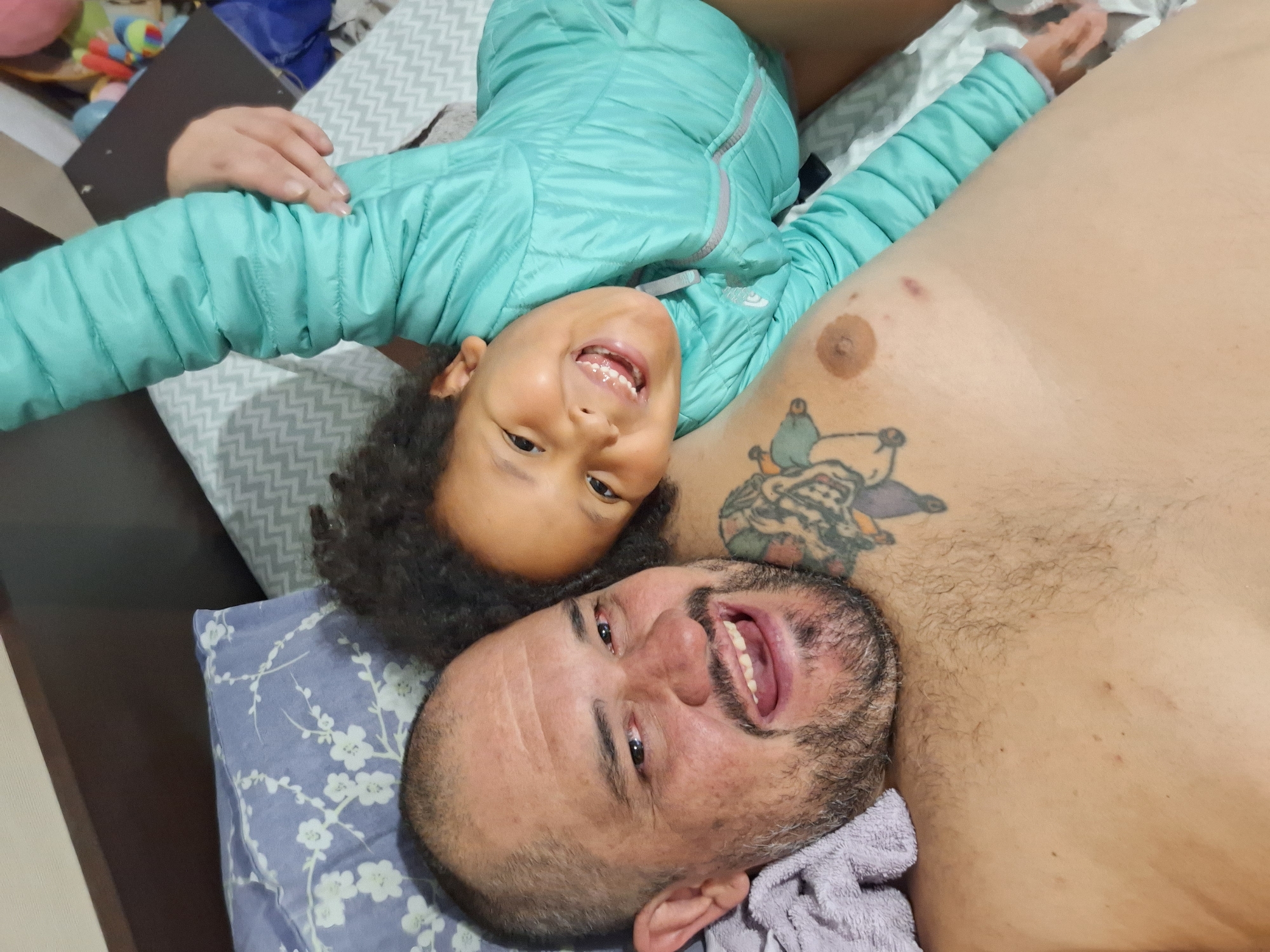 A man and a child are lying in bed, with the child resting their head on the man's chest. The man is smiling broadly at the camera, and the child is also smiling. The child is wearing a light blue puffy jacket. The man has tattoos on his chest. The bed is covered with patterned sheets and a pillow. This scene is not located in Blagoevgrad, Bulgaria.