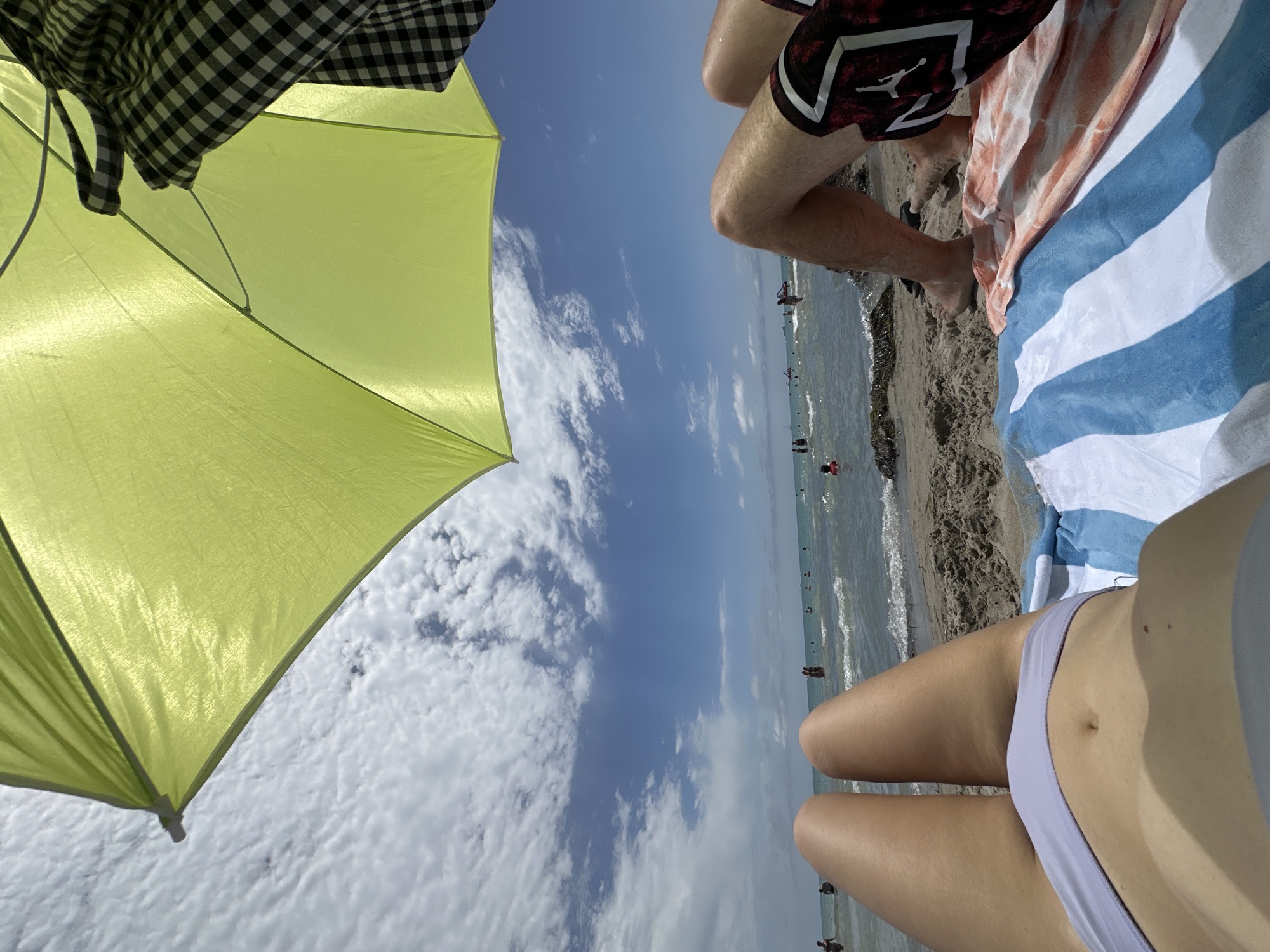 An overhead shot captures a beach scene with two individuals relaxing on towels under a bright yellow beach umbrella. The foreground features the legs of two people, one with a light purple bikini bottom and the other in patterned swim trunks. In the midground, gentle waves lap at the shore, and several people are visible in the water, some swimming and others standing. The background is a clear blue sky filled with fluffy white clouds. The location is the city of Unknown, Unknown.