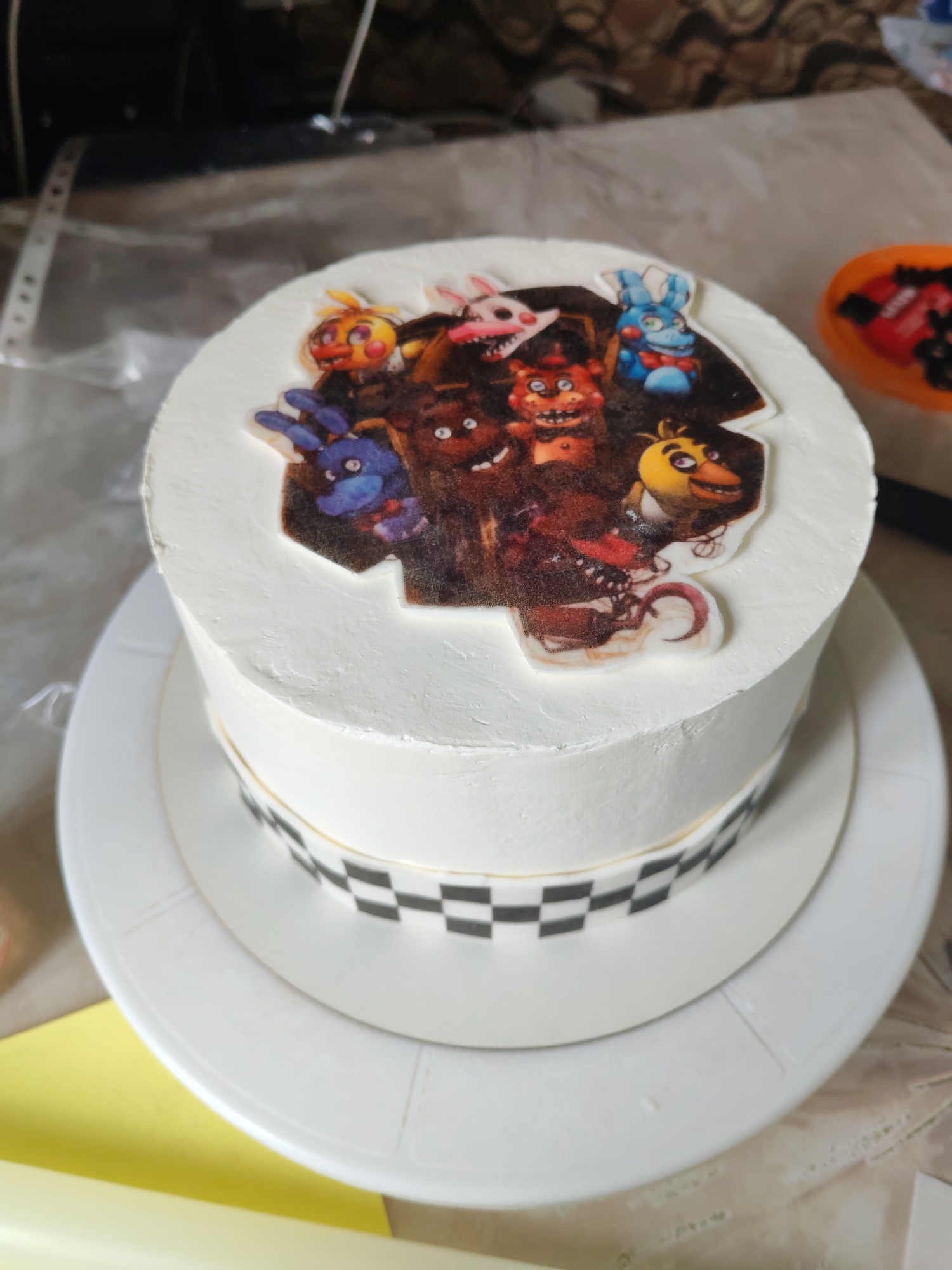 A high-angle, close-up shot presents a two-tiered cake adorned with white frosting, placed on a white serving plate. A band of alternating black and white squares encircles the lower tier. The top of the cake features a printed image depicting multiple characters from the "Five Nights at Freddy's" video game franchise. An orange container filled with dark objects is visible in the upper right corner of the frame. The background includes a table covered with a plastic sheet. The location and city are unknown.