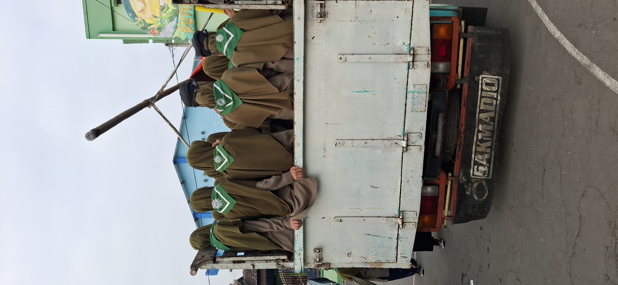 In Wonorejo, Indonesia, a group of approximately ten individuals, from a dorsal perspective, are seated in the back of a white truck. The individuals are wearing olive green hijabs and brown uniforms, each with a green scarf featuring a white emblem. Their faces are not visible. The truck is parked on a paved road with some debris scattered on the ground. In the background, a light blue building is visible, as is a green building with a mural featuring a cartoonish character. A wooden pole extends from the truck upwards, and a metal structure is attached to it. The overall scene appears to be an outdoor gathering or event.