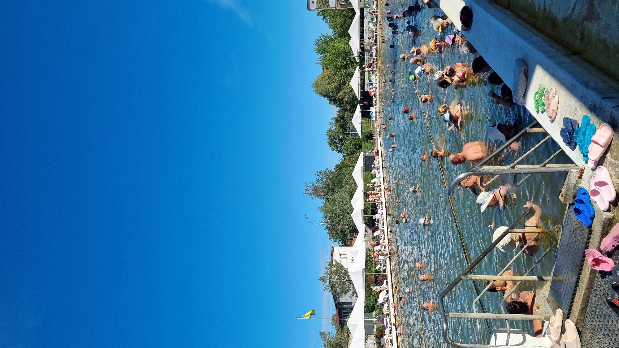 The image depicts a crowded outdoor thermal pool on a sunny day. The water is calm and murky, with numerous people of various ages swimming and relaxing. Many individuals are visible from the chest up, submerged in the water. Along the edge of the pool, there are white umbrella-like structures providing shade, with lounge chairs underneath. A yellow and green flag is visible on a pole in the background. Trees and buildings can be seen behind the umbrellas. Several pairs of flip-flops are scattered on the concrete edge of the pool in the foreground. This scene is characteristic of the thermal pools found in Berehove, Ukraine, which are known for their mineral-rich waters.