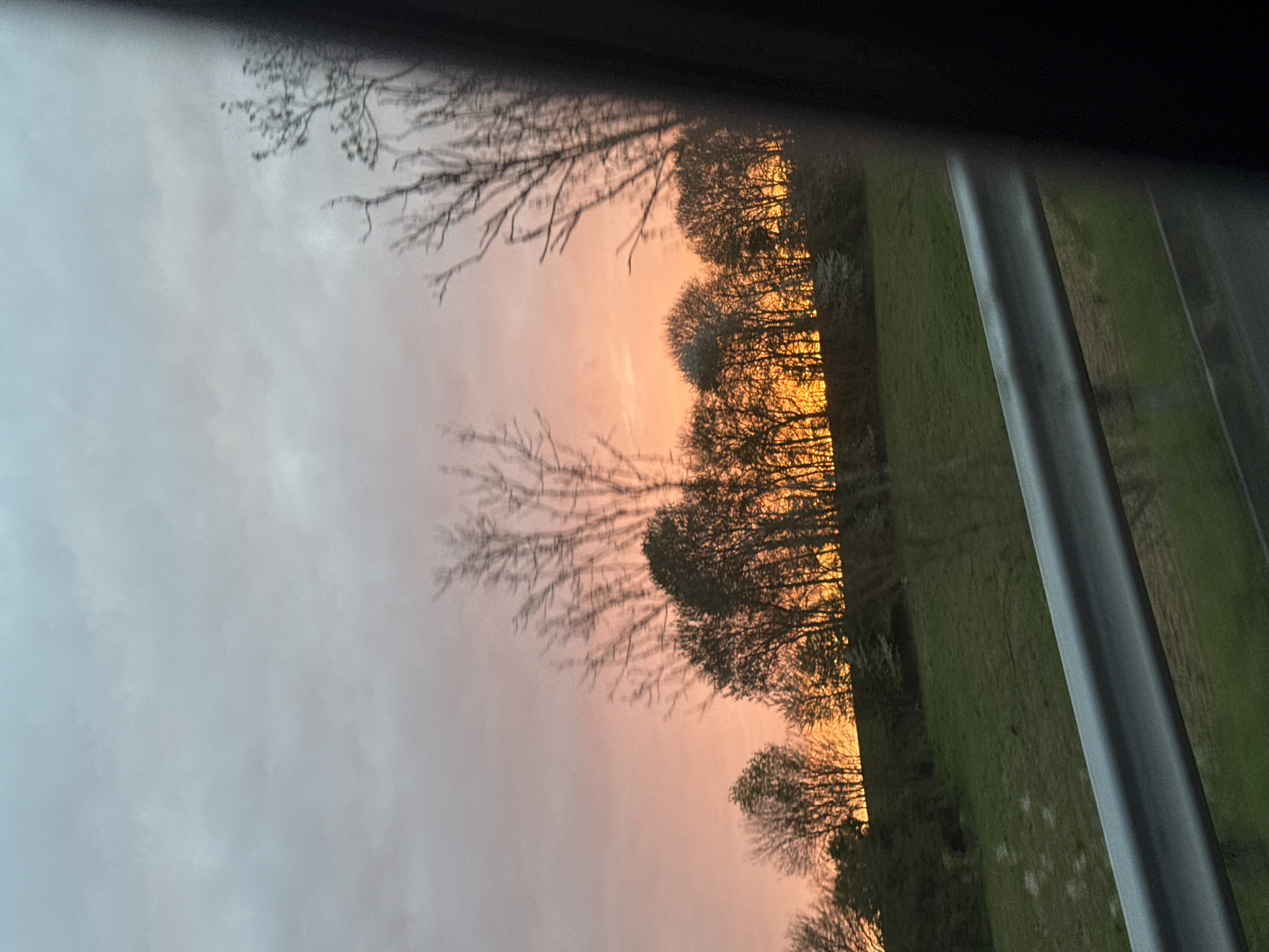 The image shows a view through a window, likely from inside a vehicle or building. The visible scene consists of a sunset sky with an orange and pink hue peeking through overcast gray clouds. Silhouetted trees are situated against the sunset. Beyond the trees, there's a green field or grassy area. A portion of the window frame is visible on the right side of the image. Without additional context, I cannot verify whether this image was taken in Saint-Quentin, France.