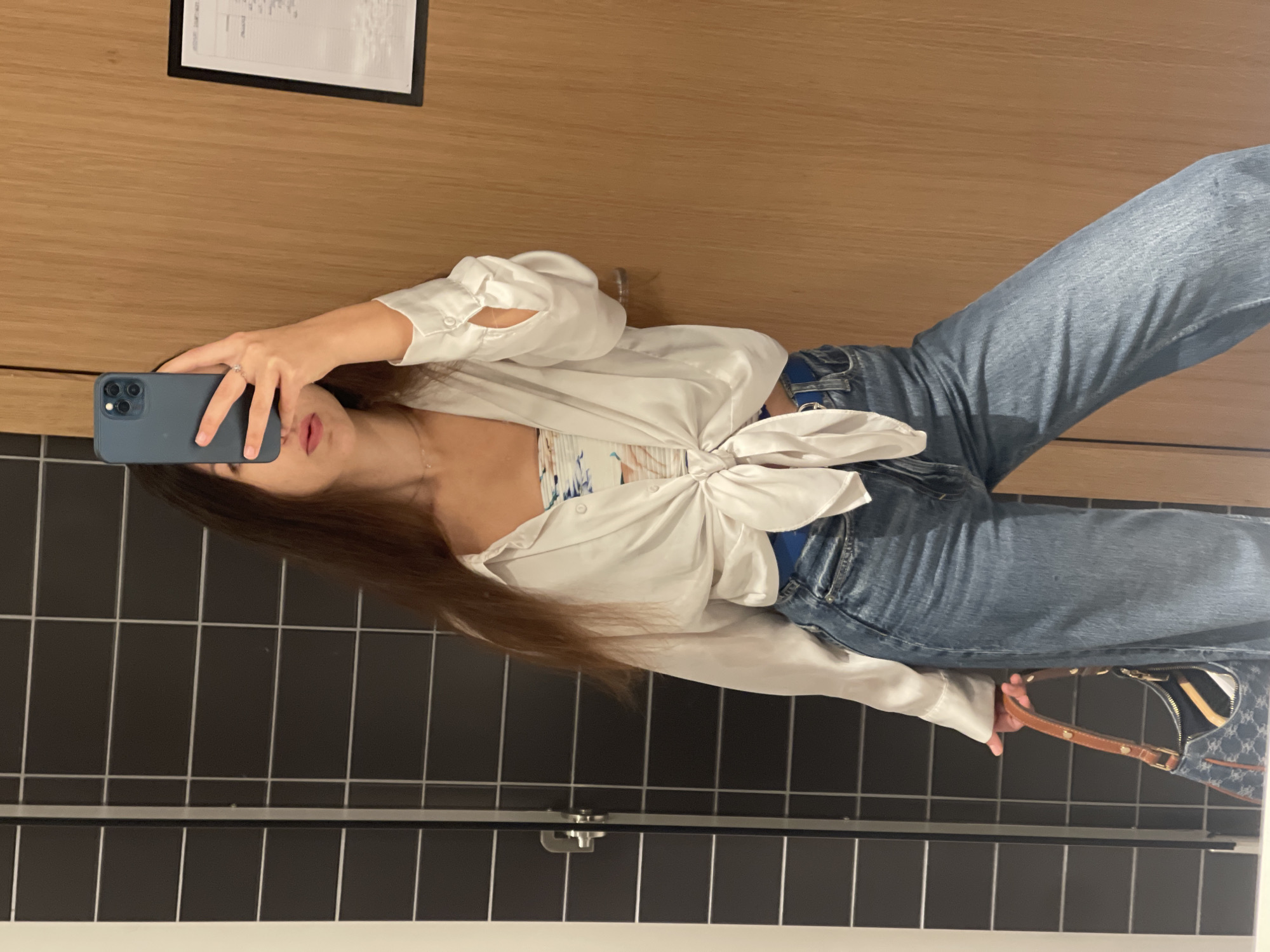 A young woman is taking a selfie in what appears to be a bathroom. She is holding a blue smartphone in her left hand, partially obscuring her face. She has long brown hair, and is wearing a white tie-front blouse over a patterned top, and blue jeans. She also has a handbag with a brown strap in her right hand. The background includes a wall with dark gray rectangular tiles and a wooden wall with a framed notice. It is not possible to determine the specific location within the city of Valencia based on the image.