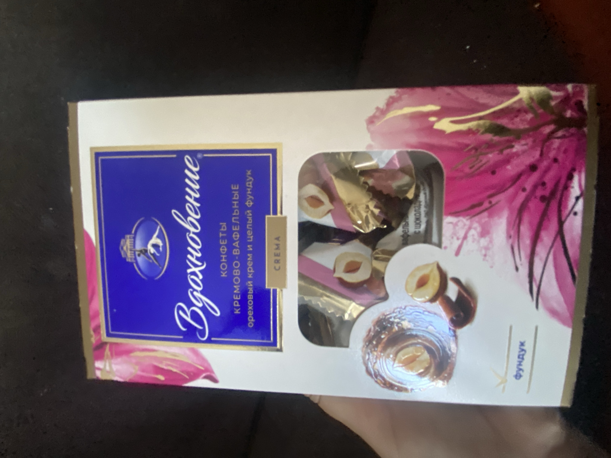 This is a product image of a box of chocolates. The box is predominantly white with blue and pink floral designs and gold accents. The brand name "Вдохновение" (Inspiration) is written in elegant script on a blue background. Below this, it states "Конфеты кремово-вафельные с ореховым кремом и целым фундуком" which translates to "Cream-waffle candies with nut cream and whole hazelnuts." There is a smaller label that reads "CREMA". The box has a window revealing several individually wrapped chocolates, some of which show a hazelnut and chocolate coating. There is also an image of a hazelnut with a chocolate swirl. On the lower left, the word "фундук" (hazelnut) is written. The background is dark and out of focus, appearing to be a textured surface, possibly a couch or blanket. The lighting suggests an indoor environment, likely a home. The image does not contain any information indicating its location in the city of Akhtyrskiy, Russia, nor does it depict any people or actions.