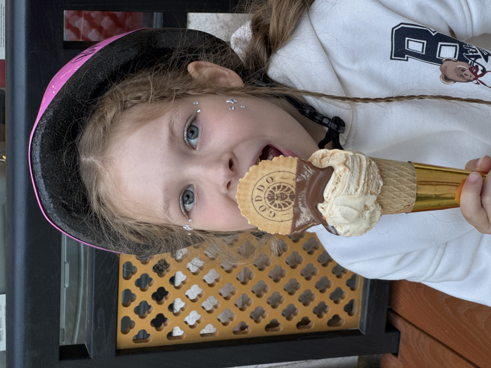 A close-up, rotated portrait shows a young girl actively eating an ice cream cone in Budva, Montenegro. The girl has light-colored hair, blue eyes, and small, reflective glitter or rhinestones adorning her face near her left eye. She wears a pink and black helmet and a white hoodie featuring a prominent black letter "P" and a small teddy bear graphic.
She is holding an ice cream cone that appears to have two flavors swirled together, one light-colored and the other coated in chocolate. A round wafer cookie, bearing the brand name "GODDO" and a crest logo, is partially embedded in the ice cream. Her mouth is open as she takes a bite from the cone.
The setting appears to be outdoors during daylight. In the background, a dark structure is visible, alongside a decorative golden-orange screen with a repeating cutout pattern resembling clovers or clouds. The girl's expression suggests enjoyment as she consumes her treat.