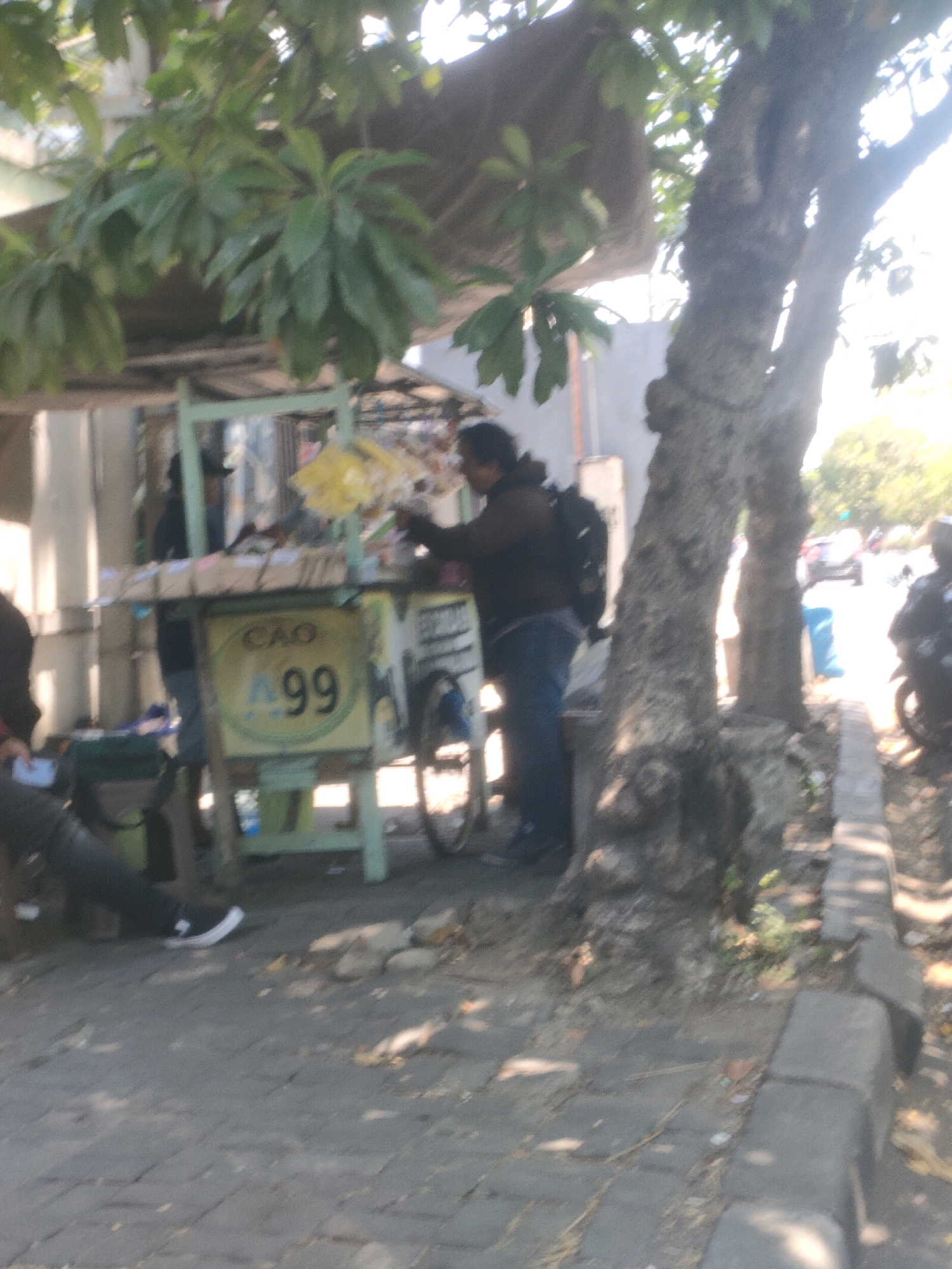 A blurred, medium-shot, outdoor scene depicts a street-side vendor stall in Surabaya, Indonesia. The stall, made of light green metal, is laden with various goods, including what appear to be bags of snacks or other items. One person, wearing a dark jacket and jeans and carrying a backpack, stands behind the stall, engaged with the merchandise. To the left, another person is seated, appearing to be holding a phone or small object. A bicycle is partially visible behind the stall. The foreground shows a paved walkway and a sidewalk with a low concrete border. A large tree with rough bark is prominent on the right side of the image, casting shadows on the ground. In the background, more stalls and buildings can be seen, along with a street with some vehicles, suggesting an urban commercial area. The lighting indicates it is daytime, with bright sunlight.