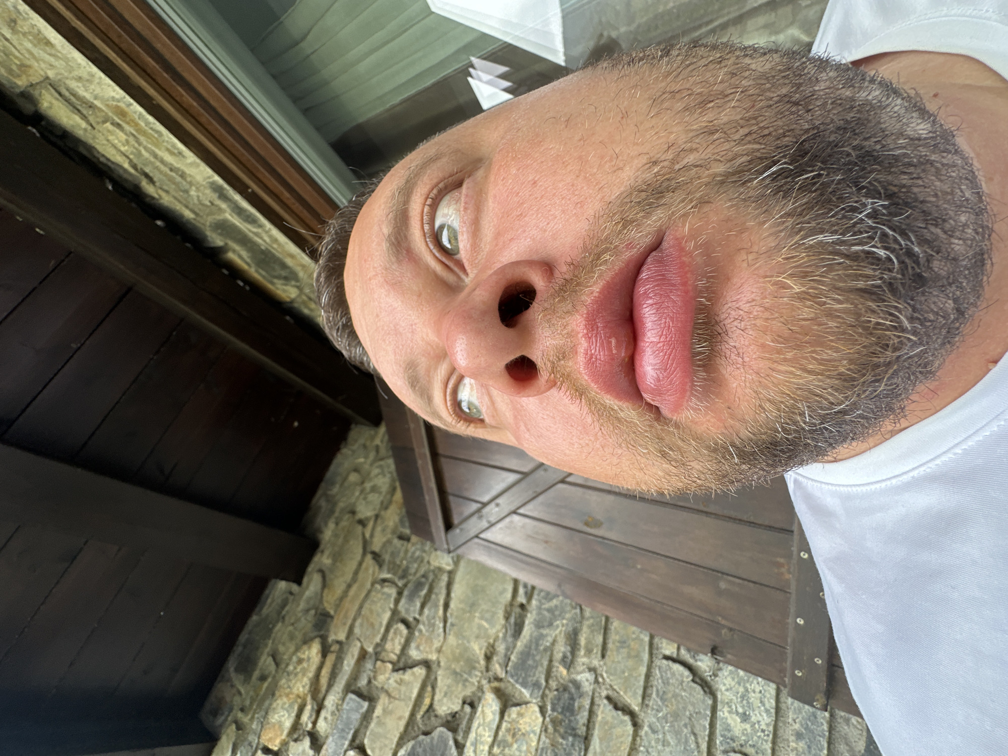 A very close-up, tilted portrait captures a man looking directly at the camera with a playful or exaggerated expression. His light-colored eyes are wide, and his lips are distinctly puckered. He has a stubble beard showing a mix of blonde, brown, and prominent grey hairs, and he is wearing a white t-shirt.
The background, also tilted, features rustic architectural elements. To the upper left and right, dark wooden planks and beams are visible, possibly part of a ceiling, eaves, or a structure. Below and to the right, a textured stone wall or pavement made of irregular, rough-hewn stones forms part of the setting. The general illumination suggests it is daytime.
The image appears to be a selfie or a close-up photograph taken outdoors or in a semi-enclosed space with natural materials. The stone and wood construction visible in the background are characteristic of traditional architecture often found in regions like Encamp, Andorra, which is the specified location context. There is no visible text in the image.