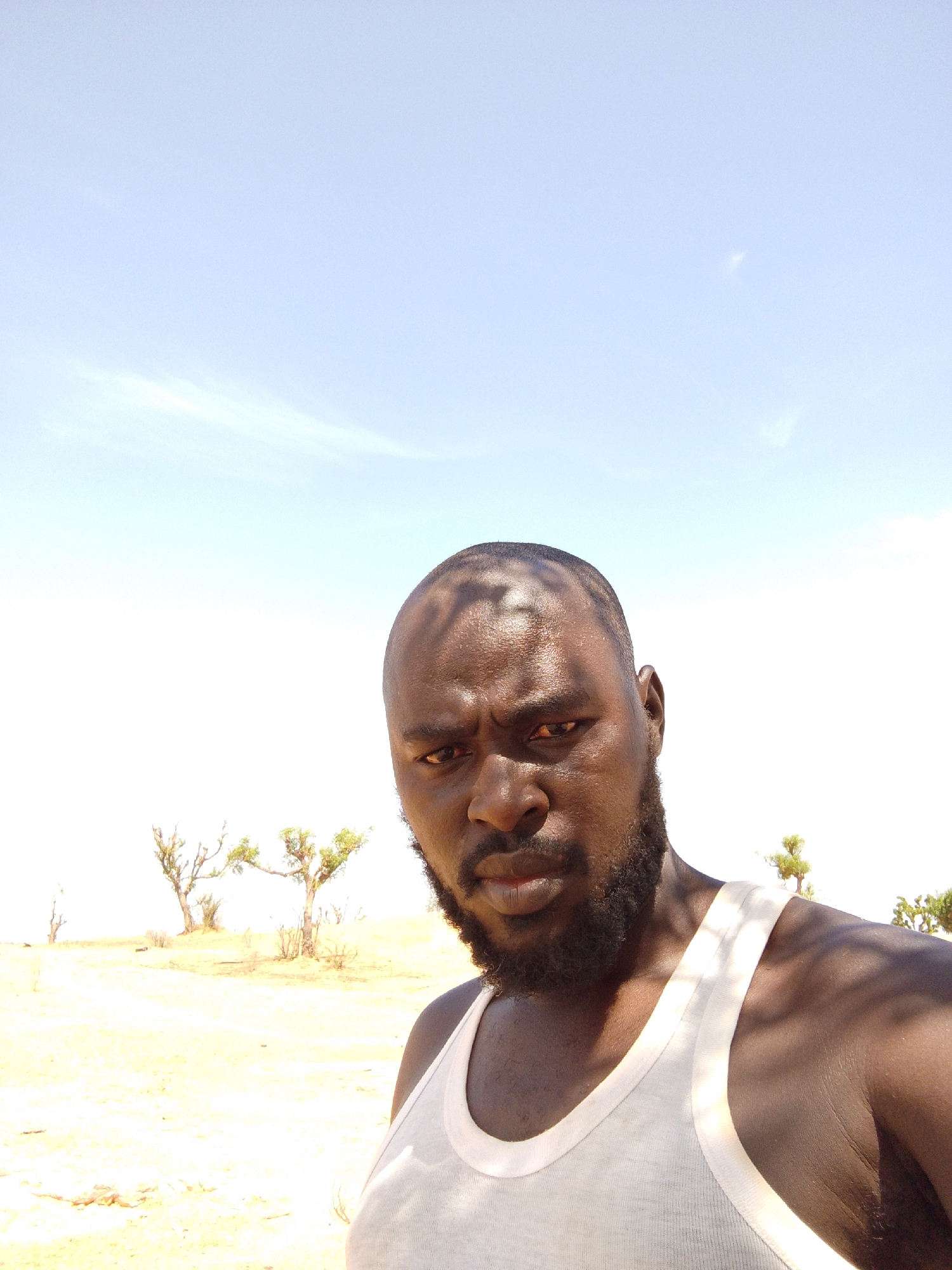 An African man in a white tank top is outdoors in a dry, sandy environment with sparse vegetation. He is facing the camera with a serious expression. The background shows a clear, bright sky and a few small trees. The location is likely Mulke Barikin Sayejo, Nigeria, based on the provided context. The photograph appears to be a self-portrait or taken at close range.