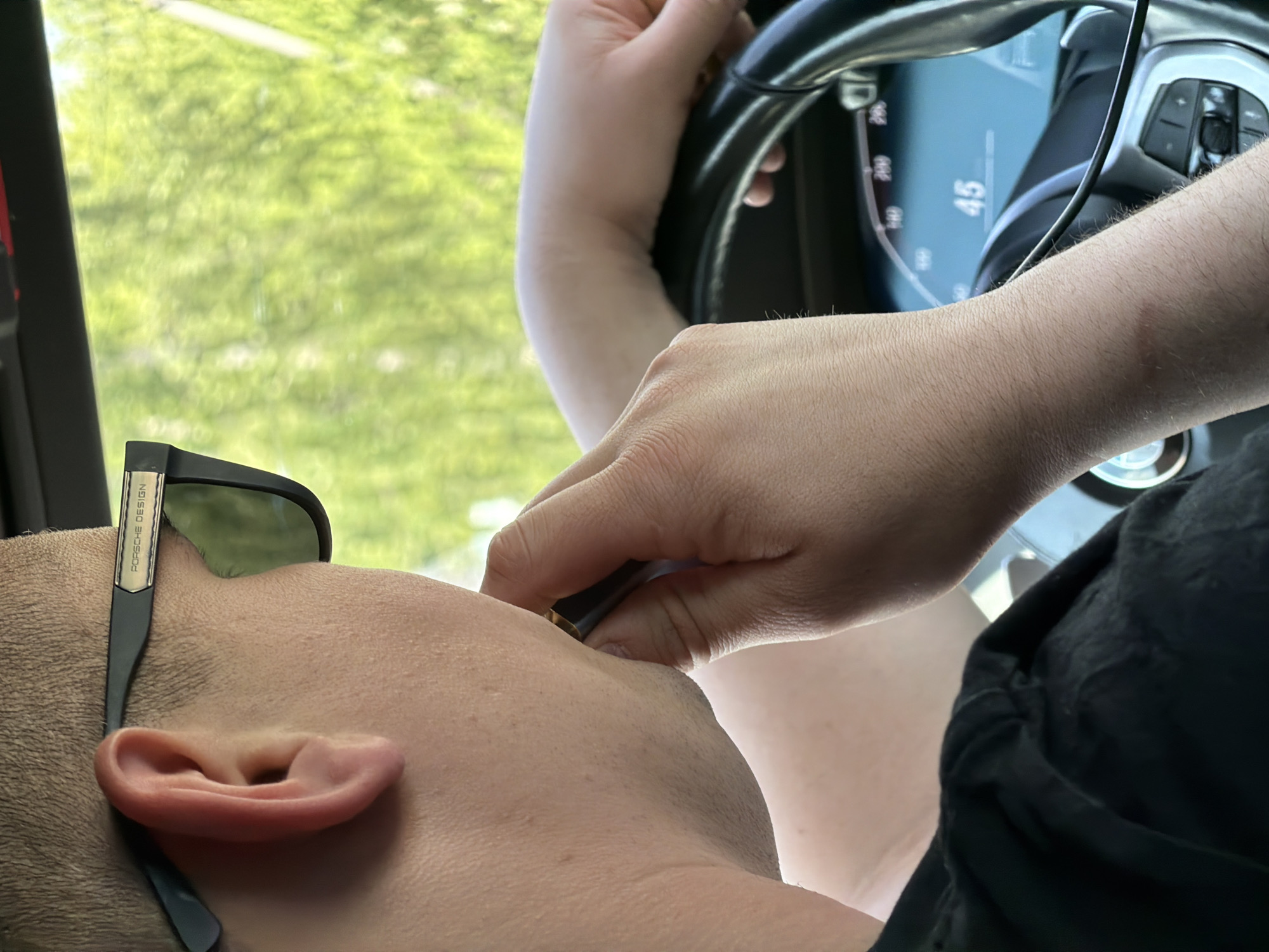 A close-up, slightly angled shot from inside a moving vehicle. A person wearing sunglasses with the logo "Porsche Design" is tilted back against the headrest. A hand is holding a small object near the person's face, possibly applying something. The driver is holding the steering wheel, and the digital speedometer reads "45". The background shows blurry green foliage through the car window. The city of Chaikovsky, Russia is not discernible from the image.
