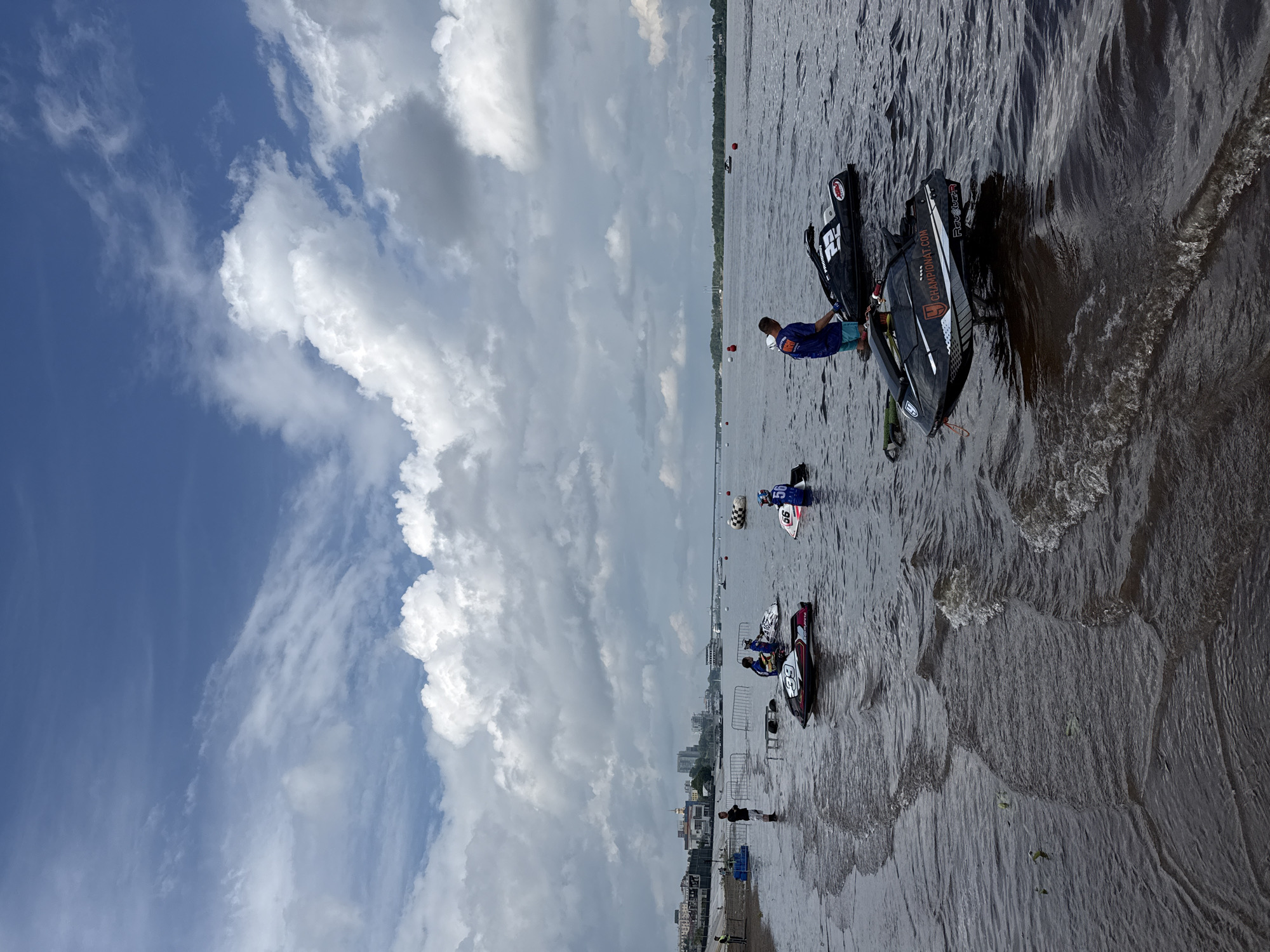 The image depicts a watercraft race event on the Kama River in Perm, Russia. In the foreground, several jet skis are partially submerged in the shallow water near the shore, some with riders or handlers present. One jet ski, black with red and white accents, prominently displays "CHAMPIONAT.COM" on its side. Further out on the water, more jet skis are visible, along with buoys that likely mark the race course. In the middle ground, a bridge spans the river, and a city skyline with buildings and cranes is visible on the left bank. The sky is filled with dramatic cumulus clouds, suggesting a dynamic weather system. The overall scene captures a moment of preparation or pause during a competitive water sports event.
