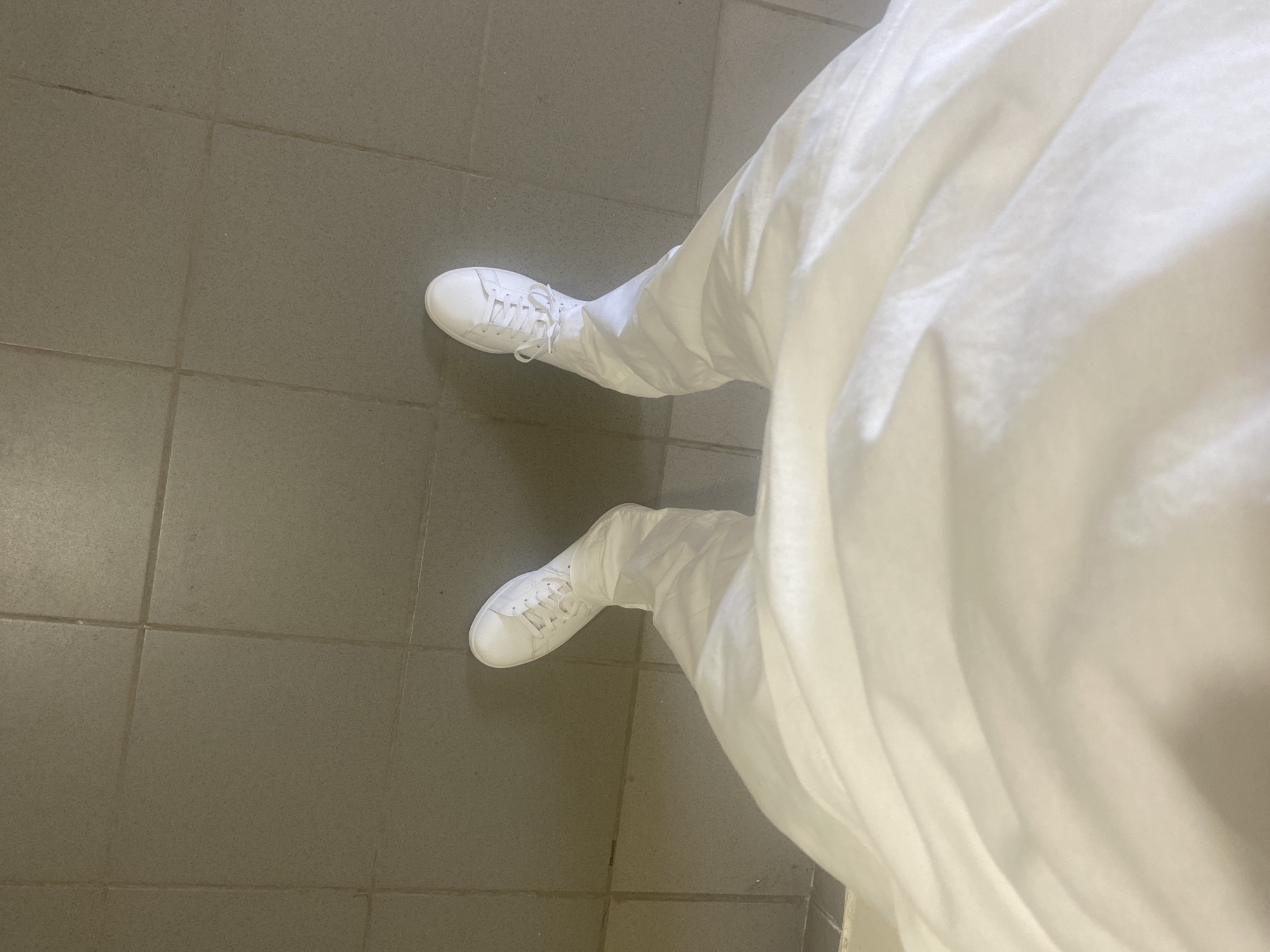 The image shows a person's legs and feet wearing white sneakers and white pants, standing on a tiled floor. The tiles are square and grey, with visible grout lines. The perspective is from above, looking down at the person's feet. There is no information available to determine the location in Novokuybyshevsk, Russia.