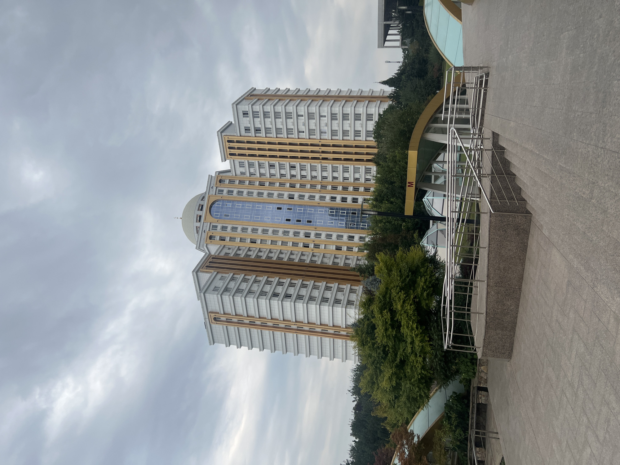The image shows a modern, multi-story residential building with a distinctive architectural design. The building is characterized by its white facade with gold accents and a prominent blue glass-covered central section. It has a domed structure at the top. In the foreground, there is a public space with paved walkways, staircases, and ramps equipped with metal railings. The area appears to be an entrance or access point to an underground facility, indicated by the "M" sign, which typically signifies a metro station. Lush green trees surround the building and the public area, suggesting a park-like setting or urban landscaping. The sky is overcast with clouds, contributing to a subdued lighting condition. The overall scene depicts a contemporary urban environment in Argun, Russia, featuring modern architecture and public infrastructure. There are no people visible in the image.