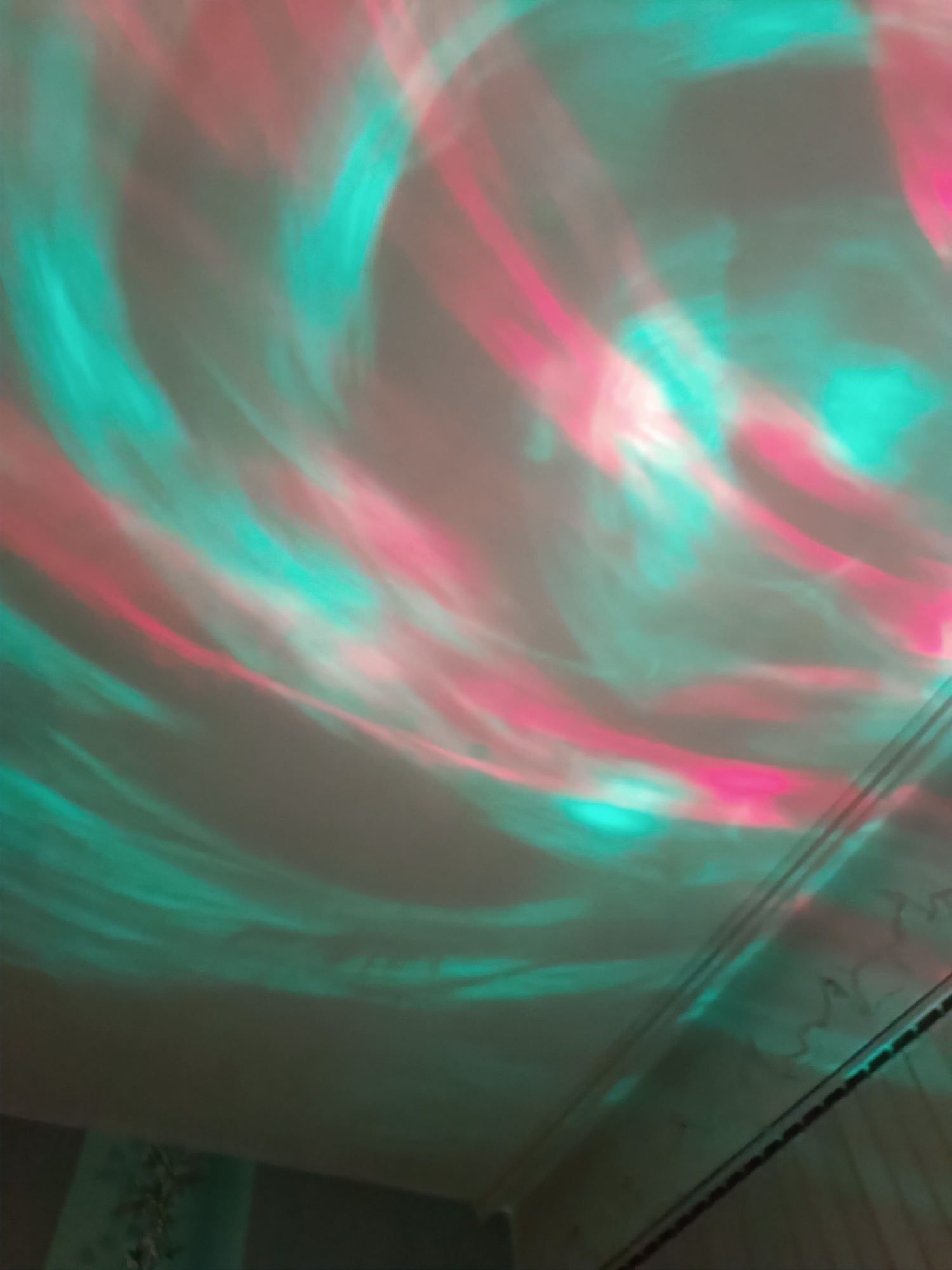 The image shows a ceiling with pink and turquoise swirling light patterns. The lower right corner reveals a section of wall with decorative trim and what appears to be a dark, possibly metal, railing or trim. The overall lighting is dim, and the focus is soft, emphasizing the light patterns. The location cannot be determined from the image alone. No information can be given about Pleven, Bulgaria.