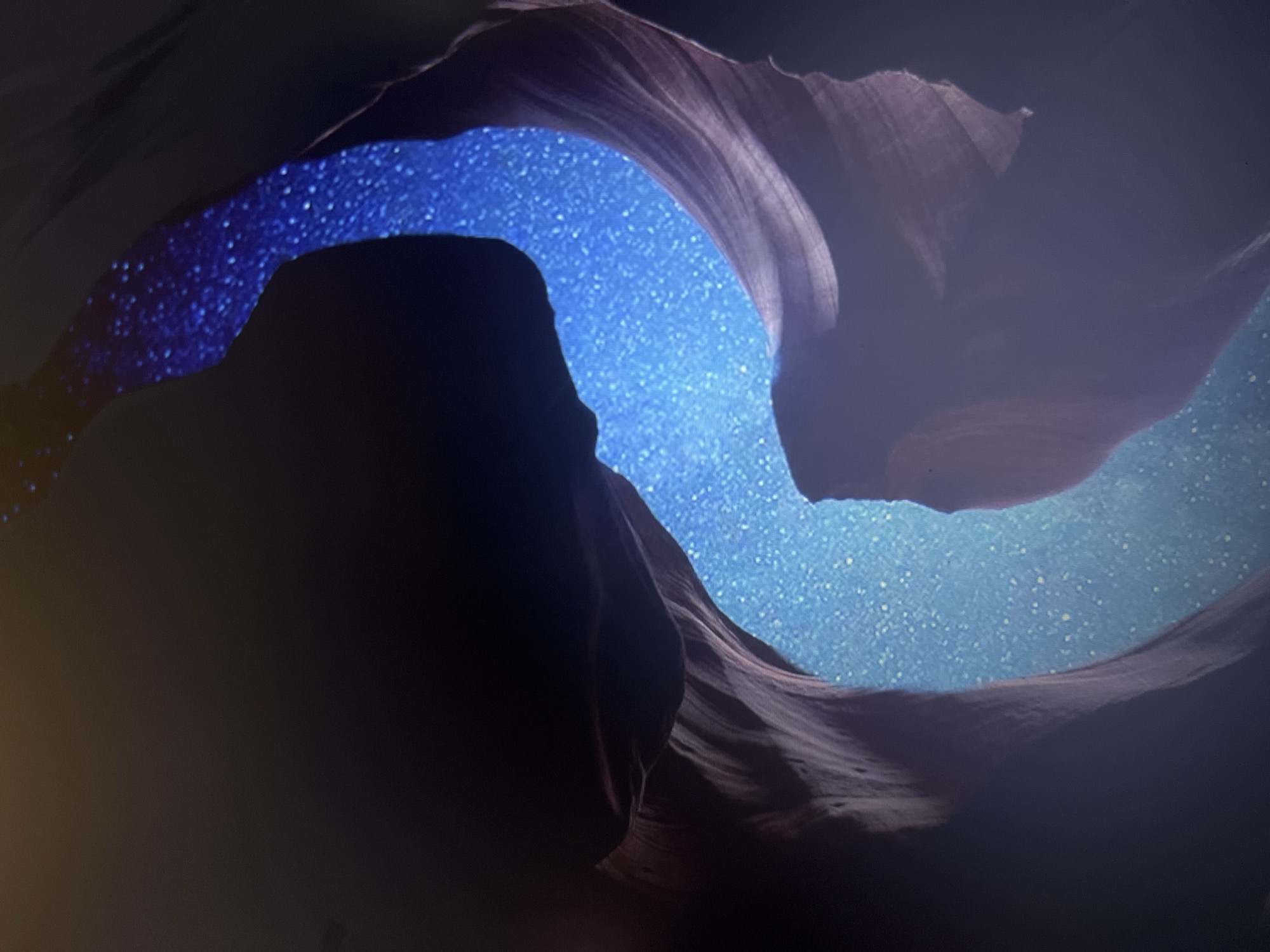 The image depicts a surreal landscape where a narrow canyon, carved from reddish-brown rock, is filled with a starry night sky instead of water or air. The canyon walls curve and twist, creating a sense of depth and otherworldly beauty. The sky within the canyon is a deep blue, densely populated with countless white and yellow stars, some appearing larger and brighter than others. The outer edges of the image are dark and shadowed, with a soft, hazy glow emanating from the starry canyon, suggesting an ethereal and dreamlike atmosphere. The overall impression is one of a natural geological formation transformed into a cosmic vista.