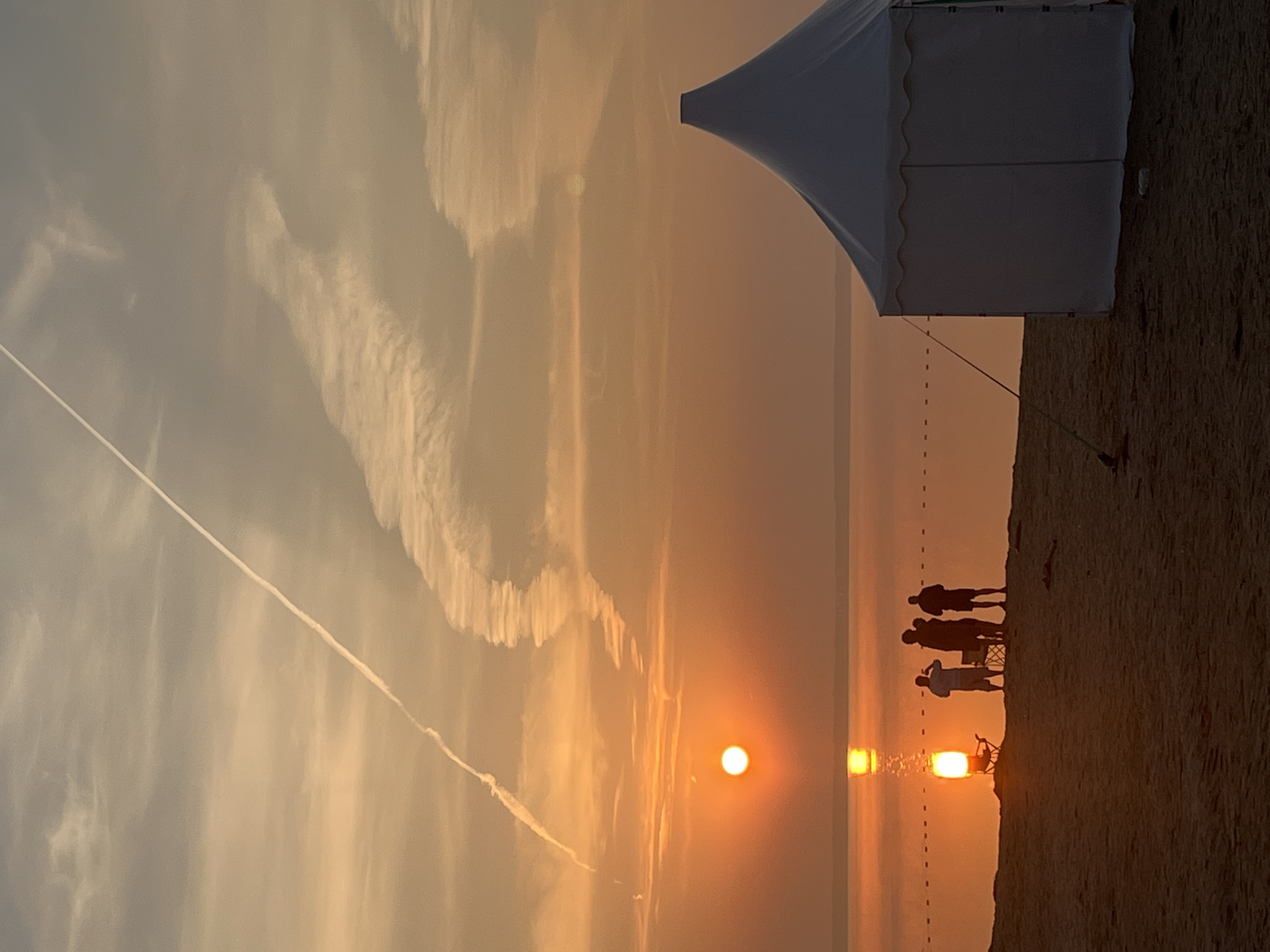 At sunset on the beach in Surakovo, Russia, the sky is painted in warm hues of orange and yellow, with wispy contrails streaking across the clouds. The sun, a bright orb, casts a golden reflection on the calm water. In the foreground, a white tent stands on the sand. A group of silhouetted figures are gathered near the water's edge, some appearing to be taking photos. One figure is seated in a foldable chair. The scene is tranquil, capturing a moment of shared enjoyment of the natural beauty.