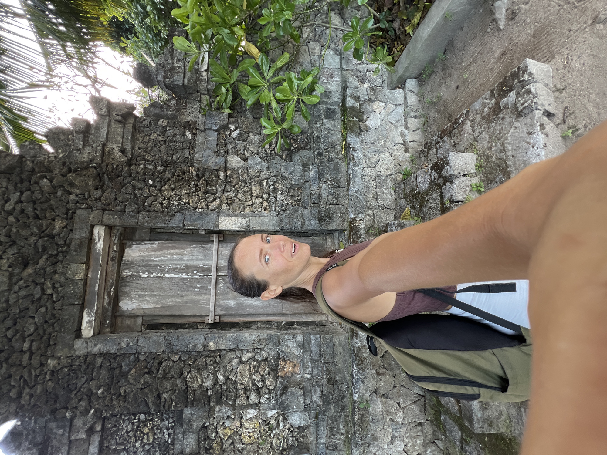 A woman stands in front of a stone wall with a wooden door. The wall appears to be part of an ancient structure. She is wearing a tank top, white pants, and a backpack. The wall has steps leading to the top. Some trees grow from the wall. The city of Denpasar, Indonesia, is not discernible in the image. The only location information is from the question prompt.