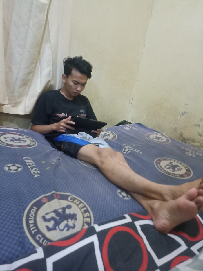 A young man, wearing a black T-shirt with the word "hulki" visible on it and blue and grey shorts, is lying on a bed with a Chelsea Football Club themed bedspread in a bedroom in Jatinangor, Indonesia. The man is holding a black tablet in his right hand and looking at it. His legs are extended and his right foot is resting on a black, red and white checkered pillow. The room has a beige wall and a white curtain. The coordinates provided, -6.934 latitude and 107.779 longitude, locate the bedroom in Jatinangor, Indonesia.