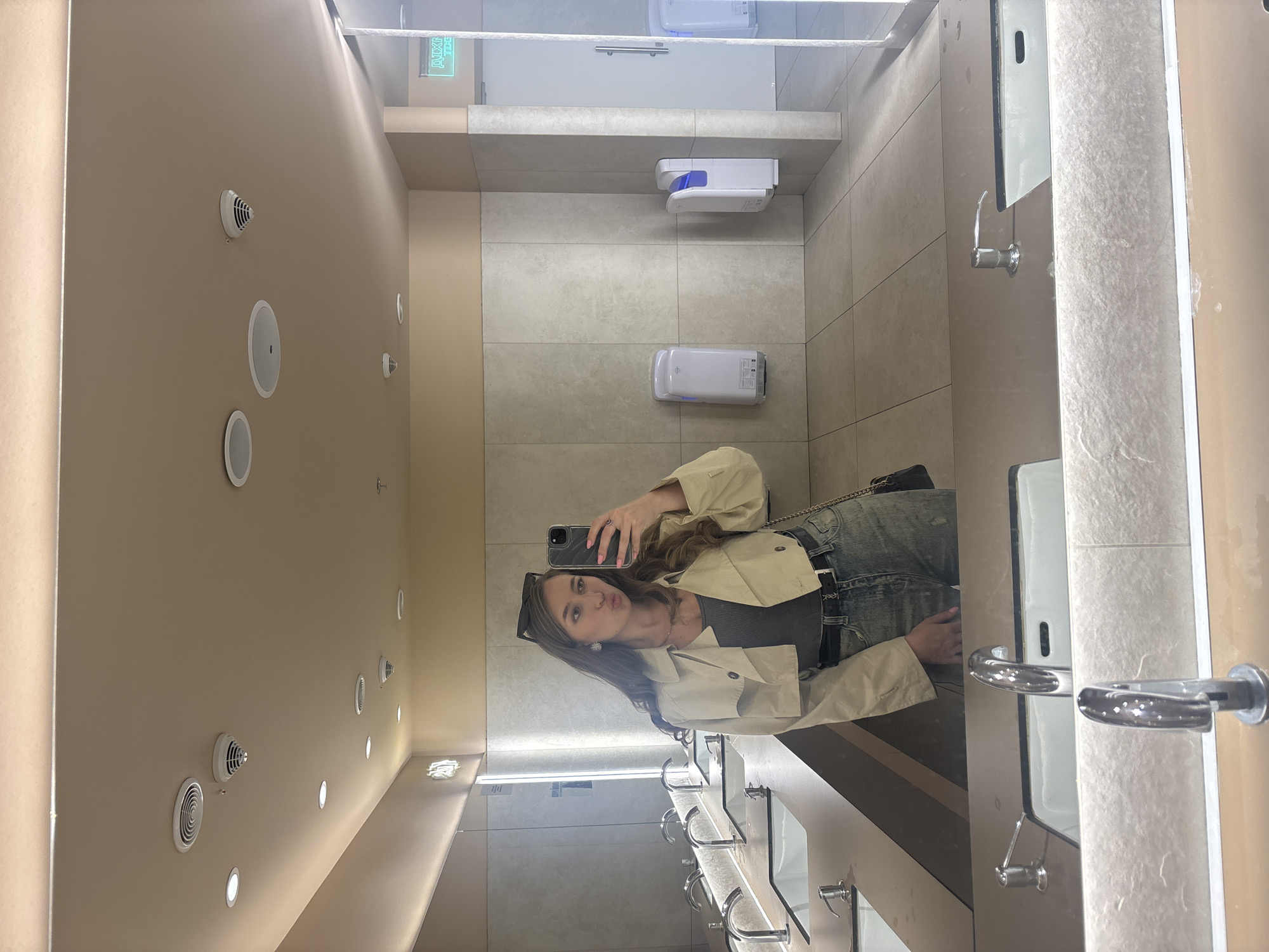 The image shows a woman taking a selfie in a public restroom. The restroom has beige walls with ventilation and lighting fixtures. Gray tiles cover the floor and sections of the walls. Soap and hand-drying dispensers are mounted on the wall above a line of sinks with mirrors. The woman is wearing a light-colored jacket, a gray top, and jeans. She is holding a phone to take the picture. The location is implied to be in Kharkiv, Ukraine, but there are no visual cues in the image to confirm this.