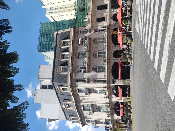 The image displays an urban street intersection on a clear, sunny day in Buenos Aires, Argentina, specifically at the corner of Calle Arroyo and Esmeralda, featuring the Palacio Estrugamou. The primary subject is a multi-story, light-colored building exhibiting Neoclassical or Beaux-Arts architecture, characterized by a dark grey Mansard roof with multiple dormer windows, ornate balconies with black iron railings, and arched windows. Its ground floor houses commercial establishments, identifiable by several dark red retractable awnings and dark glass storefronts.
Adjacent to the main building on the right, an outdoor cafe area occupies the sidewalk, equipped with multiple light-colored tables and chairs, shaded by bright red patio umbrellas. Large grey planters containing green foliage are interspersed along the sidewalk and facade.
In the background, a varied urban landscape is visible: a plain, light-colored multi-story building on the far left, a modern structure with a green-tinted glass facade situated behind the main building, and a taller, light-colored traditional high-rise further to the right.
The foreground features a dark grey asphalt road surface with a prominent white striped crosswalk (zebra crossing) extending from the lower right. A vertical three-section traffic light, a circular blue street sign, and a yellow sign are mounted on a pole on the far left. A bicycle rack with parked bicycles is also visible on the left sidewalk.
Multiple individuals are present: several patrons are seated at the outdoor cafe tables, one person walks past the main building's facade on the left, and another is partially visible near the crosswalk. The sky is bright blue with scattered white cumulus clouds. Dark green foliage from an overhead tree partially obscures the top edge of the frame.