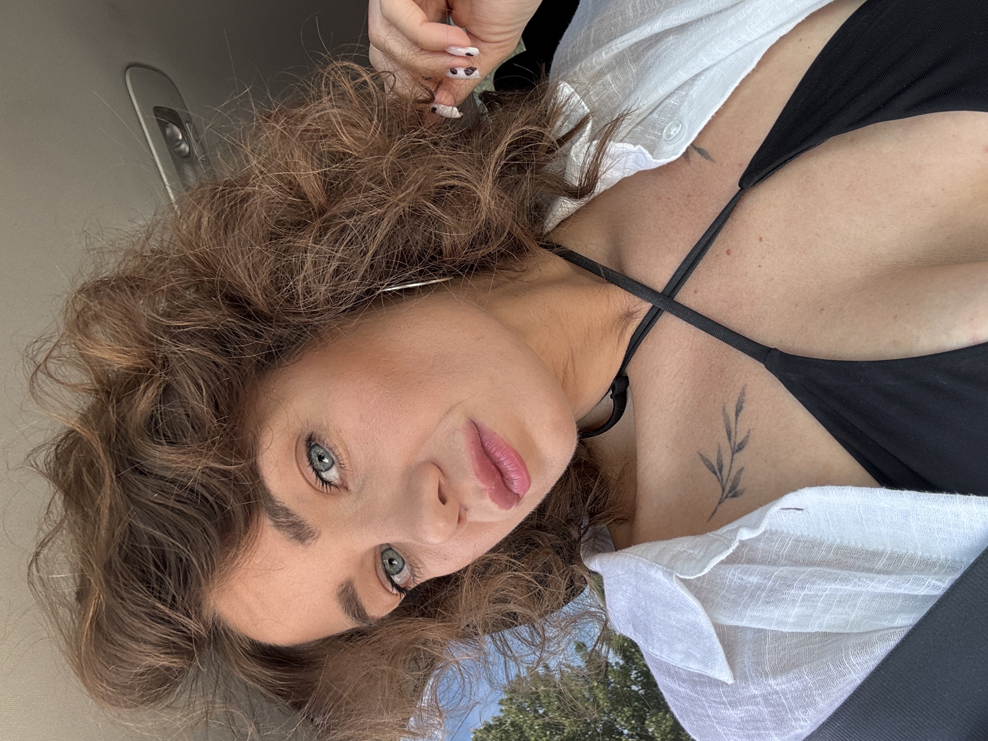 A woman with curly brown hair and blue eyes is photographed from a low angle, possibly inside a car. She is wearing a black top with thin straps and a white button-down shirt. Tattoos are visible on her chest and collarbone. The environment includes a car interior with a gray panel and a glimpse of trees and the sky outside the window. There is no information about the location in the city of Prijevor, Montenegro.