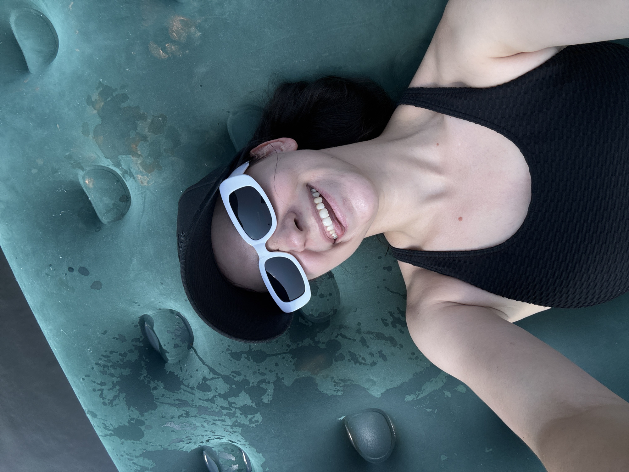 The image shows a young woman lying on a deflated inflatable mattress, which is green and has circular indentations. She is wearing a black baseball cap, white rectangular sunglasses, and a black tank top. She is smiling and sticking out her tongue. The mattress is wet and appears to be outdoors, as indicated by the sunlight. There are no visible landmarks or clues that place the location in Zasechnyi, Russia. The image is a selfie, taken from a high angle looking down at the woman.