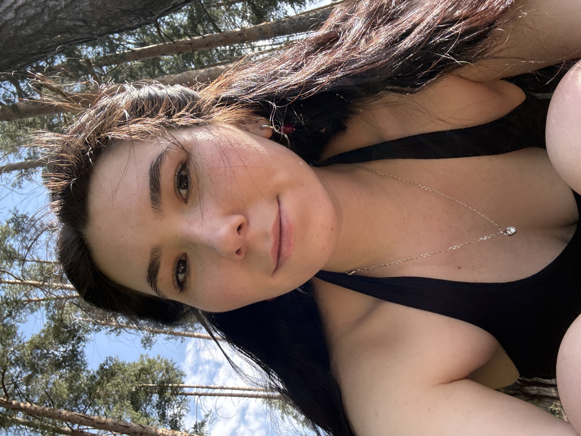 A selfie of a brunette woman with a silver necklace and black tank top is the primary subject. The background features trees and a blue sky. It is not possible to confirm the location is Izhevsk, Russia based on the image alone.
