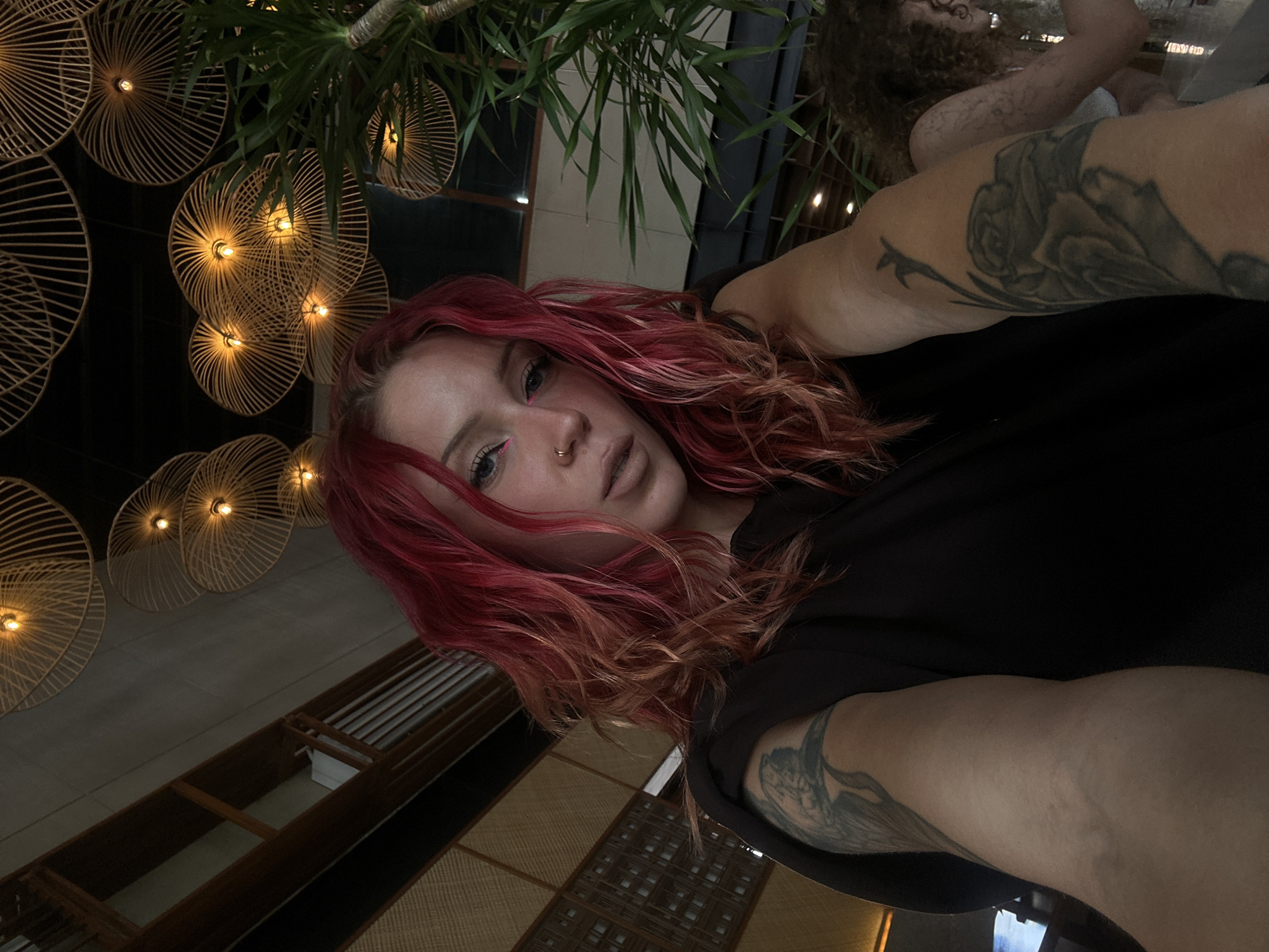 A woman with red hair and a nose piercing is taking a selfie in what appears to be a restaurant. The ceiling is decorated with round, wicker lamps and green plants. Tattoos are visible on her arms. Another person is seated in the background. The setting is likely North Kuta, Indonesia.