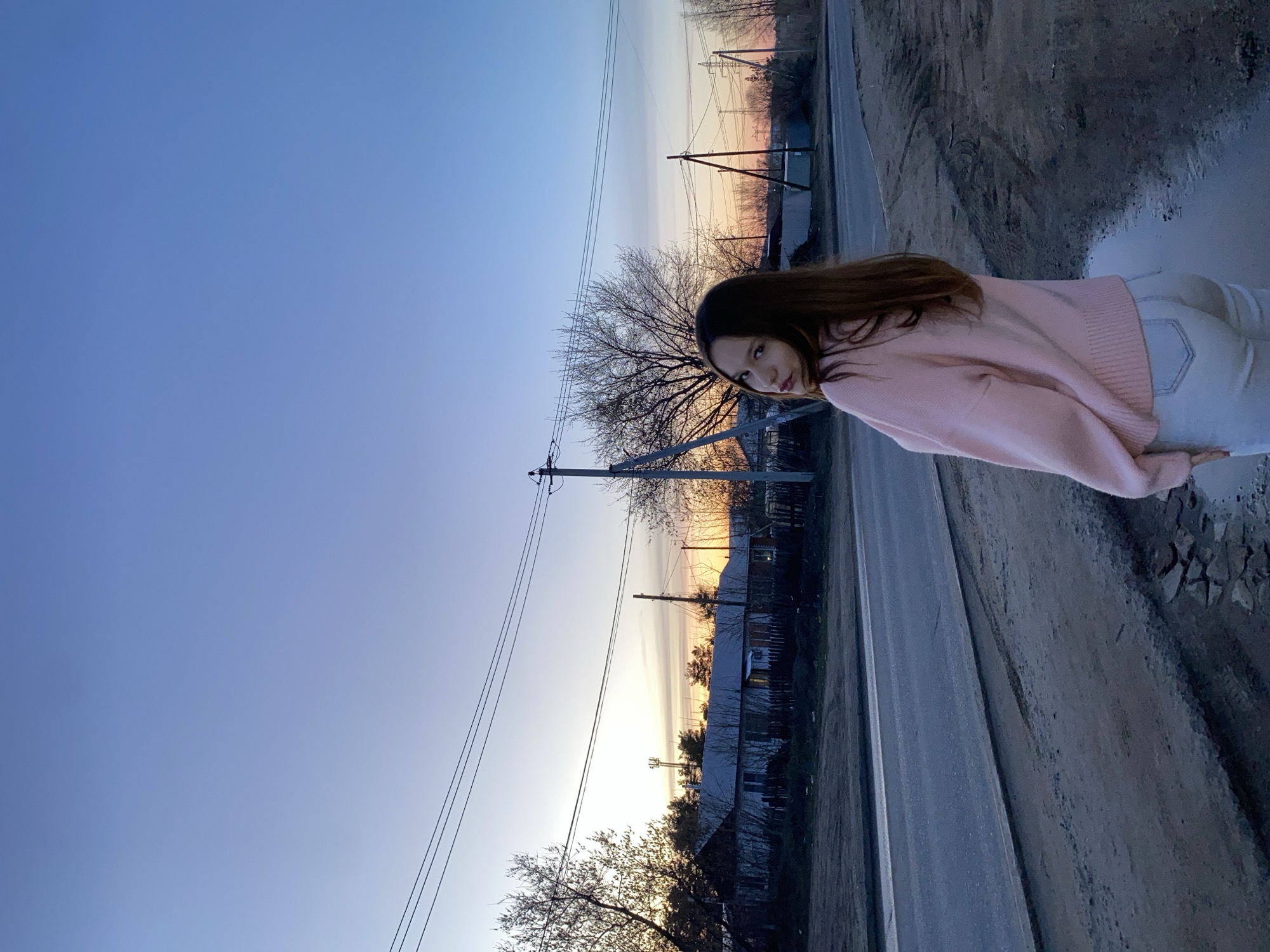 The image depicts a woman standing near a road at dusk. The location appears to be Chaikovsky, Russia, based on the presence of overhead power lines, wooden houses, and the overall landscape. The woman is wearing a pink sweater and white pants. She is standing on a dirt shoulder near a puddle. The background features a road, houses, bare trees, and a sky with hues of blue, orange, and pink.