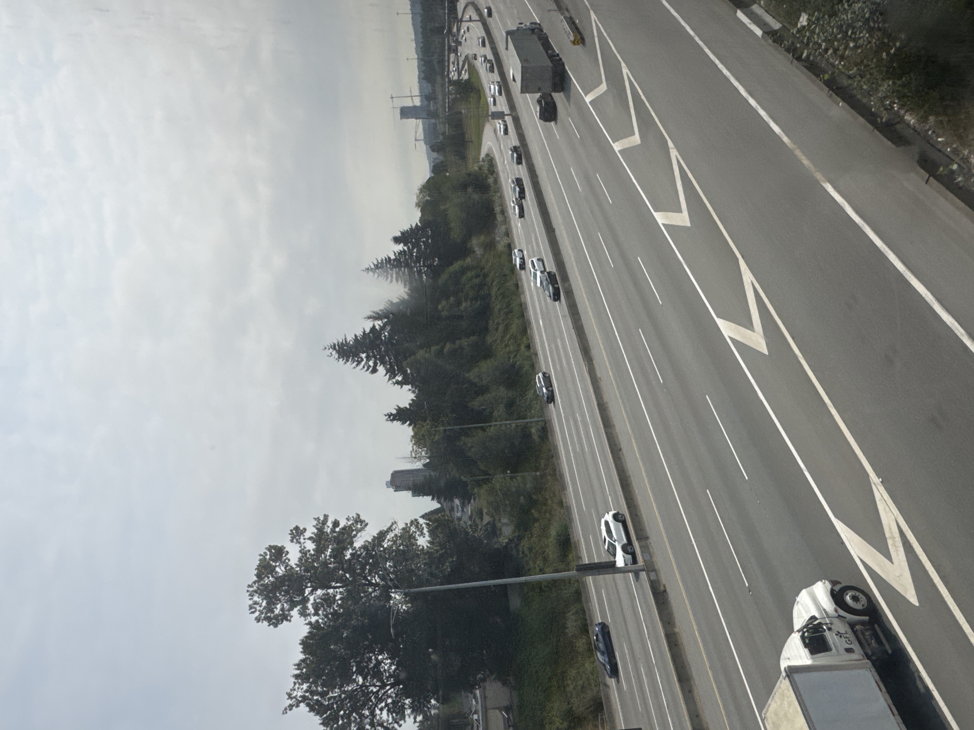 The image shows a multi-lane highway in Coquitlam, Canada, with traffic on a cloudy day. Several cars are visible on the highway, including a white SUV in the left lane and a dark sedan in the right lane of the closest visible portion of the highway. Further down the highway, more vehicles are in motion. A white truck with a box trailer is in the foreground on the leftmost lane. Trees and vegetation line the side of the highway, with a few buildings and structures, including what appears to be a high-rise building and a transmission tower, visible in the distance. The sky is overcast.