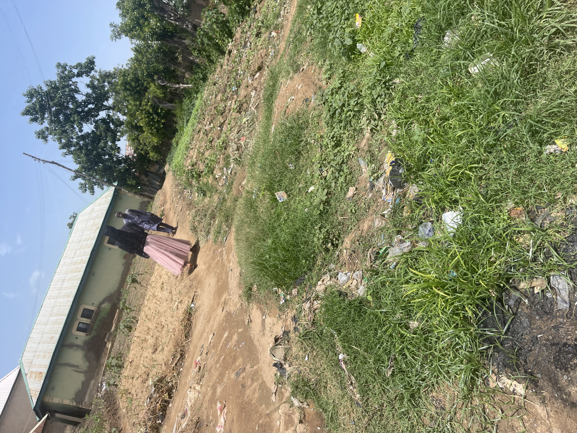 The image captures an outdoor scene in Jos, Nigeria. In the foreground, a patch of tall green grass is interspersed with scattered litter, including plastic bags and wrappers, suggesting an unkempt environment. A dirt path runs through the scene, leading towards a building in the background.
The building appears to be a single-story structure with a light green exterior and a corrugated metal roof. Two people are visible near the building. One person, dressed in a dark outfit with a black headscarf, wears a long, pale pink skirt. The other person, standing slightly behind and to the side, is dressed in a dark patterned outfit. They appear to be walking or standing together.
The ground around the building and path is dry and dusty, with more scattered debris. Trees with lush green foliage are visible behind the building and to the left, indicating a vegetated area. The sky above is a clear, bright blue, suggesting it is daytime, likely a sunny day. The overall impression is a residential or semi-rural setting with elements of neglect due to the visible litter. There is no visible text in the image.