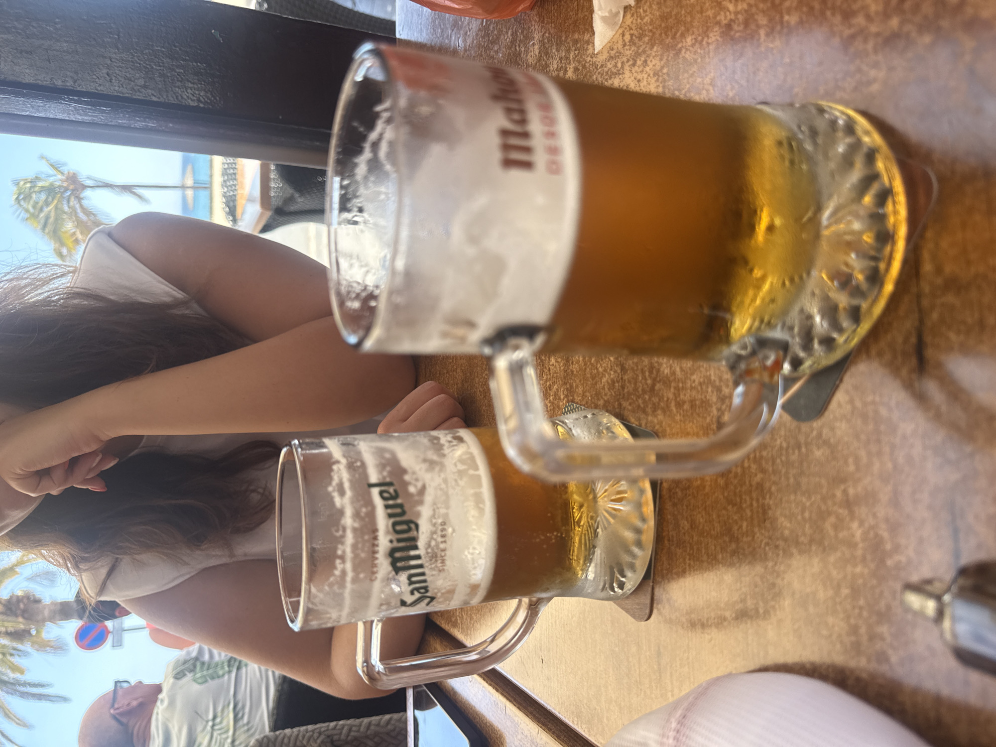 The image depicts a vibrant outdoor scene from a cafe or restaurant, likely in a sunny, coastal location such as Puerto Del Carmen, Spain. The view is from a seated perspective, looking across a table towards the sea.
In the foreground, two large, handled beer mugs rest on a light brown, possibly wooden, table surface. The upper mug, closer to the camera and partially consumed, clearly displays the "Mahou" brand name on its label, featuring a substantial head of foam. Below it, a fuller mug is visible, branded "San Miguel" with "CERVEZAS SINCE 1890" printed beneath, also topped with a foamy head. Both beers are light amber lagers.
Two individuals are partially visible. On the left side, the arm and part of the upper body of a person with long, dark hair and a white top are seen, resting on the table. Further back and to the left, the profile of a bald man wearing glasses and a light-colored, patterned short-sleeve shirt (reminiscent of a Hawaiian shirt) is visible, looking towards the background.
The background is dominated by clear blue skies and bright sunlight. Several lush palm trees are prominent, suggesting a tropical or subtropical environment. Beyond the trees, a sparkling blue expanse of ocean or sea is visible. A red and white circular road sign, possibly a "no entry" or "stop" sign, can be seen amidst the greenery. The bright, clear weather and setting evoke a relaxed holiday atmosphere, suggesting daytime enjoyment of drinks by the sea.