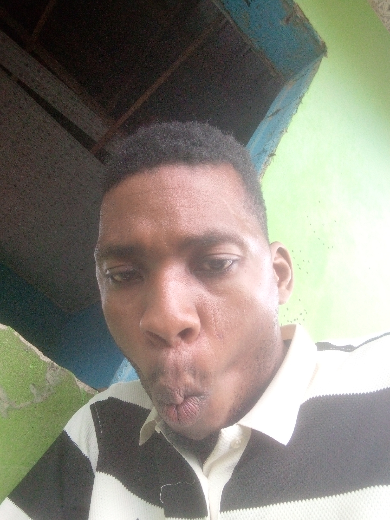 A man with short, dark hair and brown skin is shown from a close, upward angle. His lips are pursed in a kissing or blowing motion. He is wearing a polo shirt with alternating black and white horizontal stripes and a white collar. Behind him, a green wall is visible on the right and lower left, and above him is a dark, likely interior ceiling with exposed wood beams and decaying blue paint. The location appears to be indoors, though the condition of the walls and ceiling suggests a poorly maintained or aged structure. It is not possible to confirm the location is Ikorodu, Nigeria without additional information.