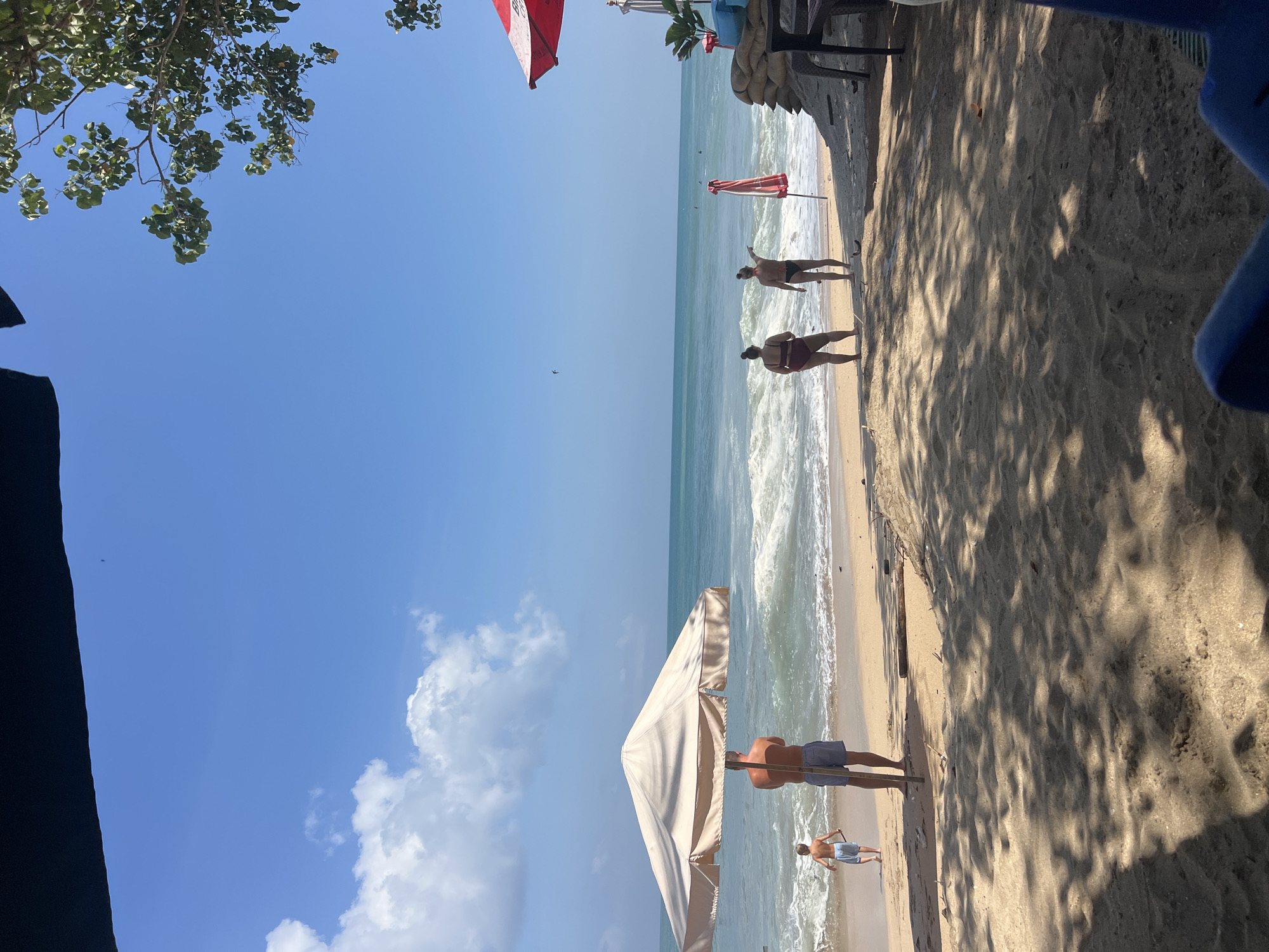 The image depicts a beach scene in Kuta, Indonesia, on a clear, sunny day. The foreground is a sandy beach with dappled shade from overhead foliage. In the mid-ground, several people are present. A man in blue shorts stands under a large, beige beach umbrella, facing away from the camera towards the ocean. To his left, a young boy in light blue shorts is walking towards the water. Further down the beach, two women in swimsuits are standing near the shoreline. One woman, wearing a dark red bikini, is facing the ocean, while the other, in a black bikini, is pointing towards the water. A red and white beach umbrella is planted in the sand near them, along with sandbags forming a low barrier. The ocean has moderate waves breaking on the shore, and several surfers can be seen in the distance. The sky is bright blue with scattered white clouds. The overall atmosphere is one of leisure and a typical beach day.