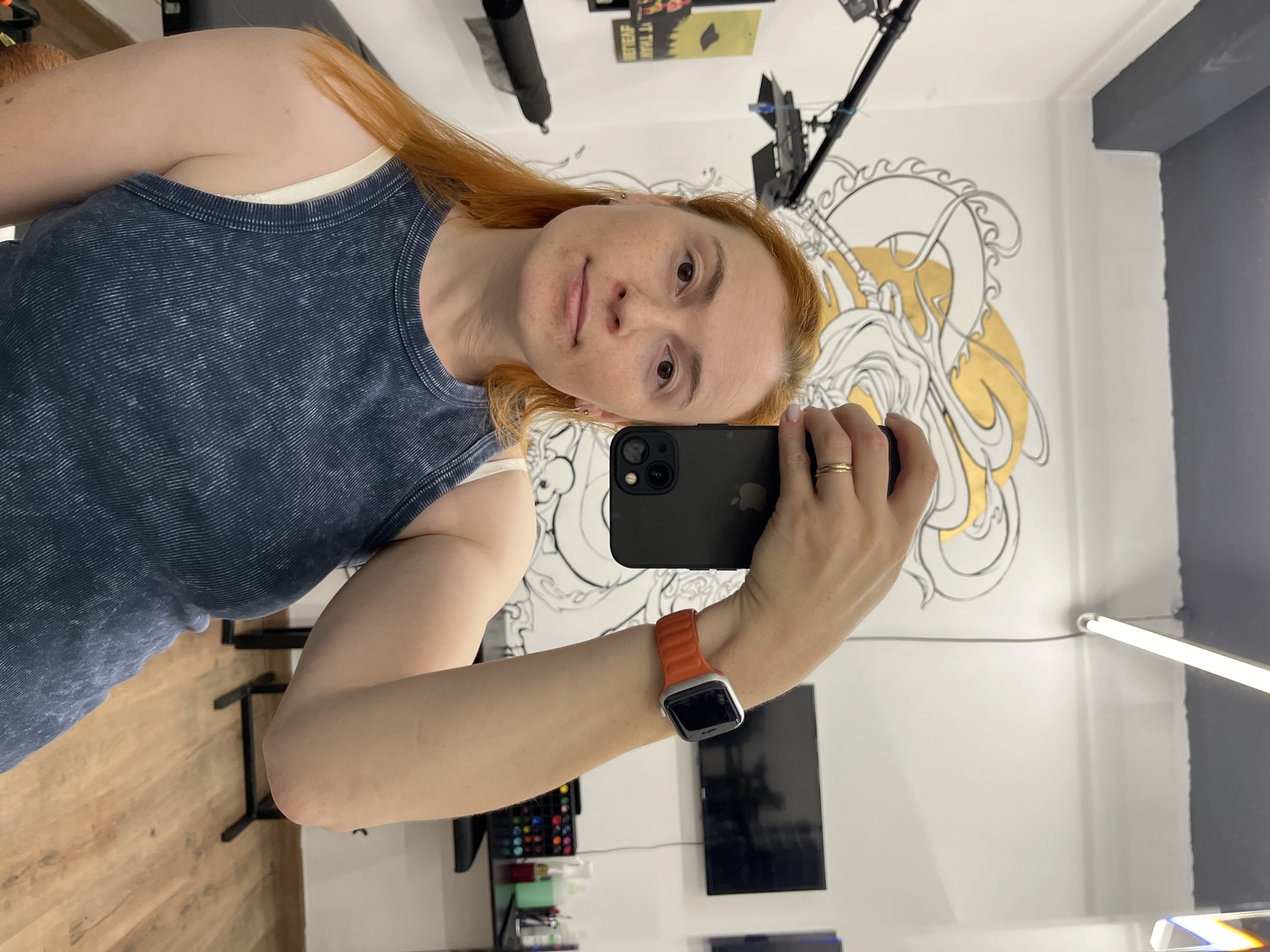 A young woman with long, reddish-blonde hair and fair skin is taking a selfie in what appears to be a tattoo parlor in Chaikovsky, Russia. She is wearing a dark blue, ribbed tank top over a cream-colored bra. Her left arm is raised, holding a black smartphone. On her left wrist, she wears an orange Apple Watch band. The background features a wall with a large black and gold mural, a TV screen, a rack of colorful markers, and various tattoo equipment.
