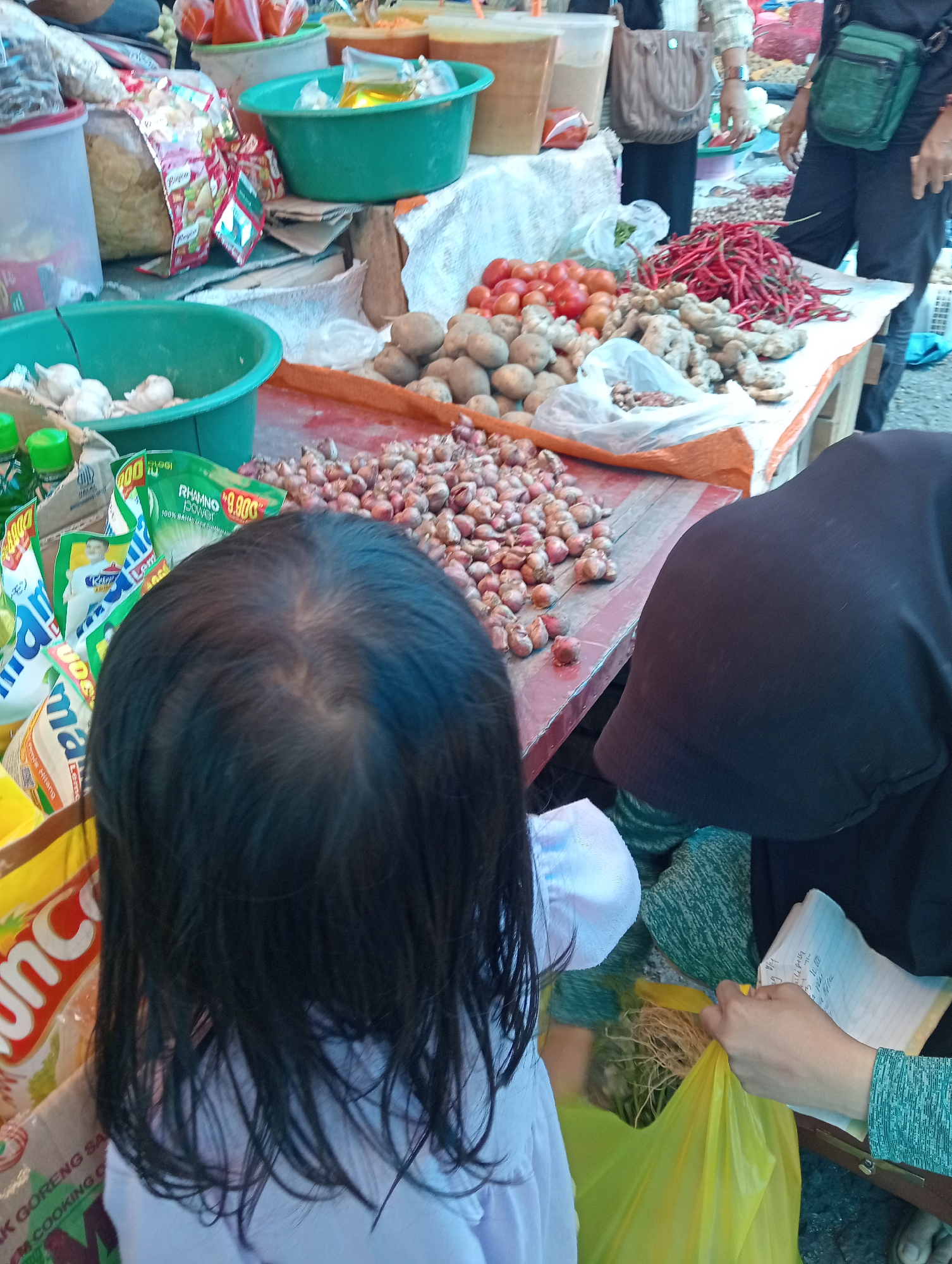 This is a street market scene in Pariaman, Indonesia. A young girl with dark hair is facing away from the camera, wearing a light purple shirt. To her right, a person wearing a black hijab and a teal and black patterned shirt is holding a yellow plastic bag filled with green onions or herbs. The person's hand, wearing a teal and black patterned sleeve, is reaching into the bag. Another hand, also with a teal and black patterned sleeve, is holding a small notepad with writing on it. The market stall displays a variety of produce, including piles of small red onions, potatoes, tomatoes, and long red chilies. There are also green basins containing garlic and other items, as well as packaged goods with Indonesian text. The ground is paved and appears to be asphalt.