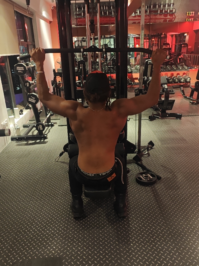 A male individual with a muscular physique, bare-chested and wearing black athletic pants, a black backward baseball cap, and black athletic shoes, is seated facing away from the viewer on a black lat pulldown machine. His hands firmly grip a straight bar extended upwards, indicating a paused or extended phase of the exercise. A silver-colored chain is visible around his neck, and a light-colored band is on his left wrist. The waistband of black underwear with white lettering ("SPORT" and a "CK" style logo) is visible above his pants. The gym floor is covered with dark grey diamond-patterned rubber matting. The background features various exercise equipment, including racks of dumbbells with "HAMMER STRENGTH" branding (e.g., 22LB weights visible), stacked weight plates, and other machines, all reflected in large wall mirrors. A loose 10kg weight plate lies on the floor to the right, and a white industrial fan is visible on the left. The gym is illuminated by artificial red and white lighting, creating a distinct ambiance in this fitness facility located in the Chongqing High-tech Zone, China.