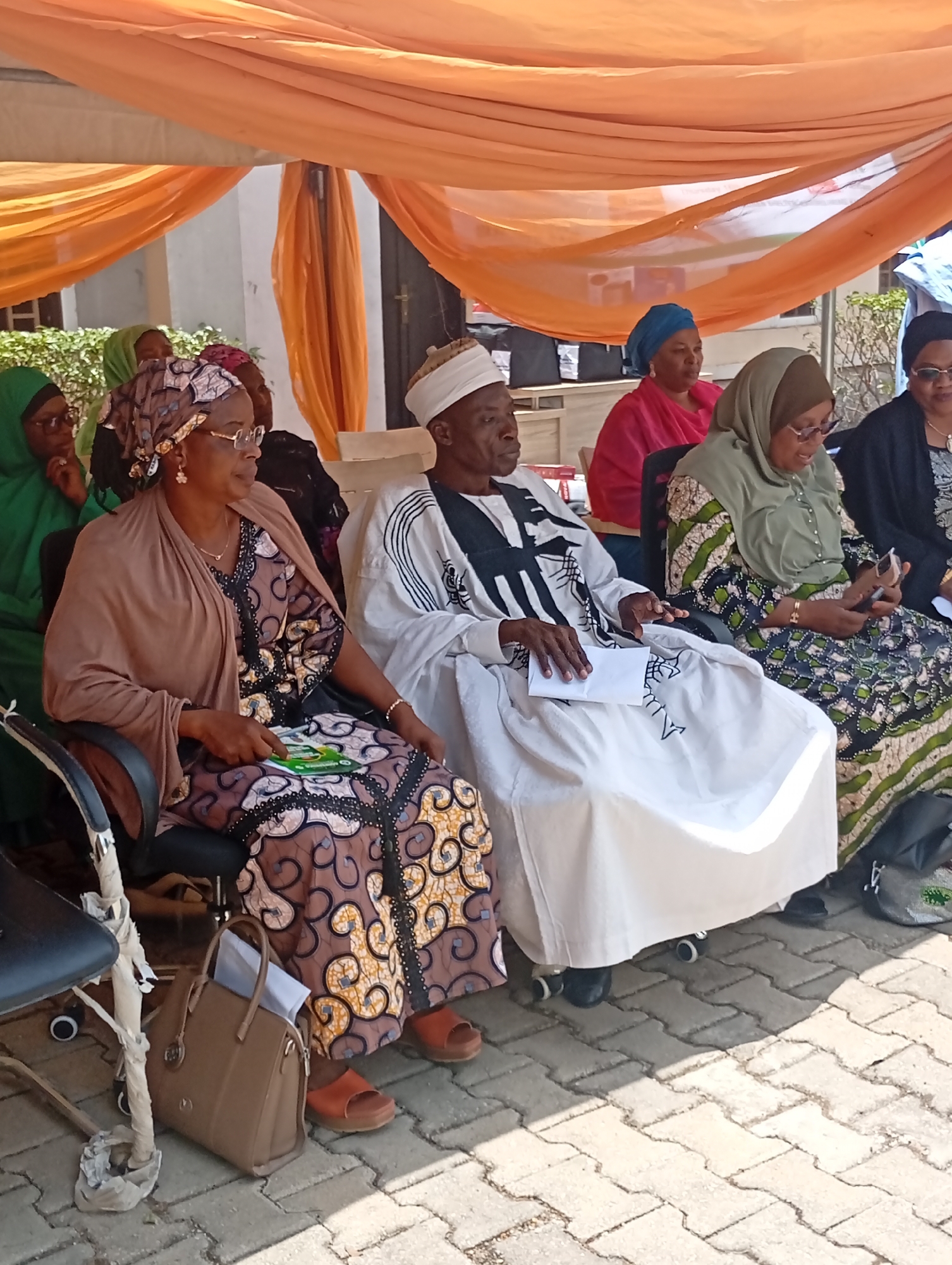 This image depicts an outdoor gathering taking place in Unguwar Sarki, Nigeria, under bright, sunny daylight, evidenced by distinct shadows cast on the paved ground. The event is held beneath a large orange canopy, which provides shade.
A group of approximately seven to eight adults are seated in a row of black chairs. In the foreground, a woman wearing a brown and intricately patterned long dress, a matching headscarf, and a light brown shawl is seated on the left. She wears glasses and holds a green leaflet that clearly displays the word "KOKO" in white lettering. A brown handbag rests on the ground beside her chair.
Central to the visible group is an older man, dressed in a traditional long white gown adorned with distinctive black stylized patterns, and a white cap. He holds a piece of white paper in his hands, resting them on his lap. To his right, another woman in a light olive-green hijab and a patterned dress holds and looks at a mobile phone. Further down the row, partially visible, are more attendees, including a woman in a bright pink top and a blue headscarf.
The participants appear attentive and calm, suggesting a formal or semi-formal meeting, workshop, or community event. The background includes part of a light-colored building and some green foliage, indicating an open-air or courtyard setting. The attire of the individuals aligns with traditional fashion often seen in the region.