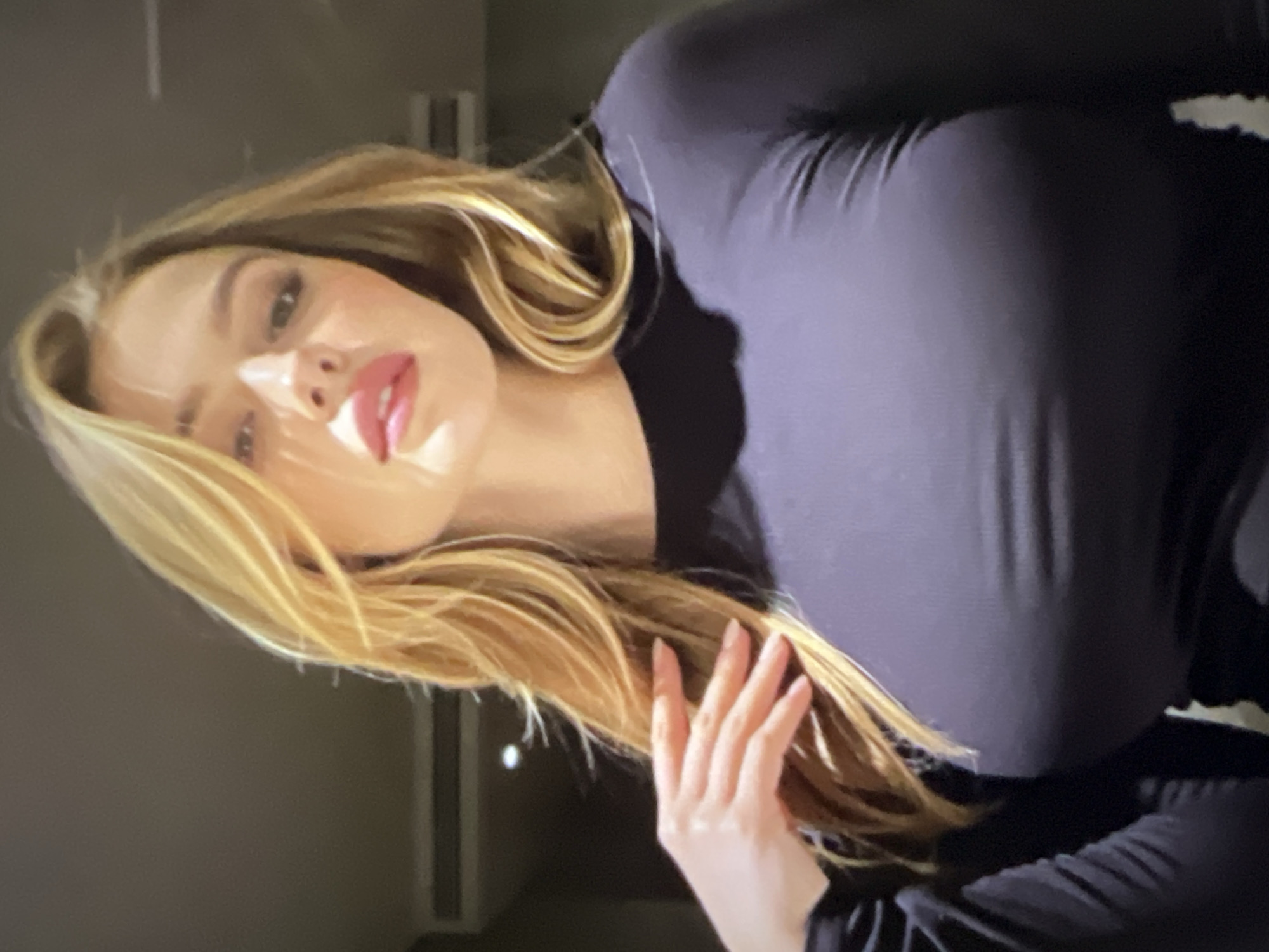 This media file shows a close-up, vertically oriented, medium shot of a young woman. She has long, blonde hair that is slightly tousled and falls around her shoulders and chest. Her skin is fair and appears smooth. She is wearing a dark, form-fitting top that accentuates her figure. Her head is tilted to her right, and her gaze is directed towards the viewer. She has prominent makeup, with bright pink lipstick and defined eyelashes. A soft light illuminates the left side of her face, creating a strong highlight and casting a shadow on the right. Her left hand is visible in the foreground, near her chest, with fingers slightly spread. The background is a plain, dark, and out-of-focus surface, possibly a wall or a piece of furniture, with some vertical elements that are indistinct. There is no visible text in the image. The overall impression is one of a posed or candid portrait, with a focus on the subject's facial features and overall aesthetic. The lighting suggests an indoor setting, possibly with artificial light.
