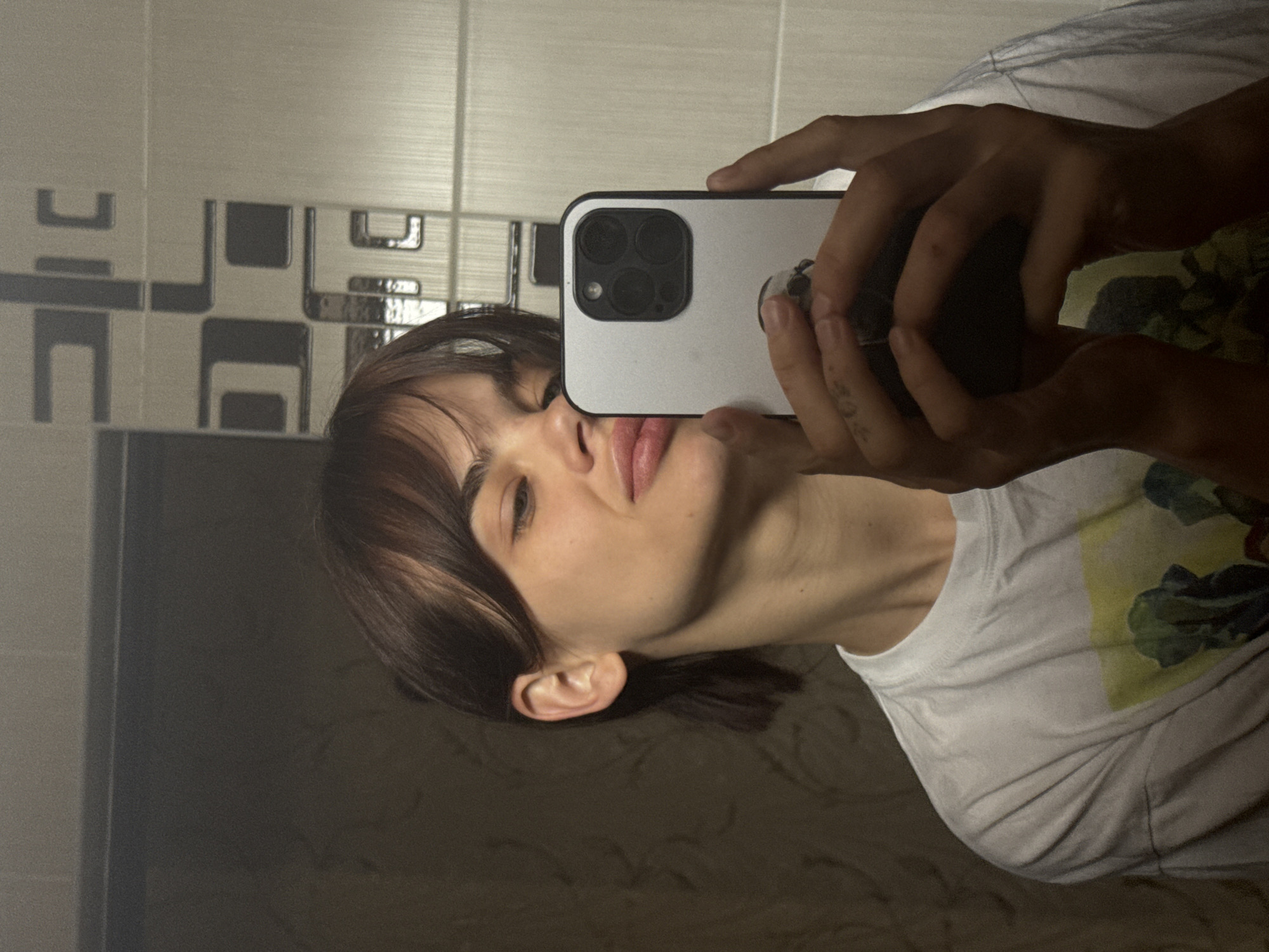 A person is taking a selfie in front of a tiled wall and mirror. The person is holding a white iPhone with a multiple lens camera. The person has brown hair with bangs and is wearing a white t-shirt. The tiles on the wall behind the person are geometric patterns of black and white. The location is not identifiable as Rostov-on-Don, Russia, from the image alone.