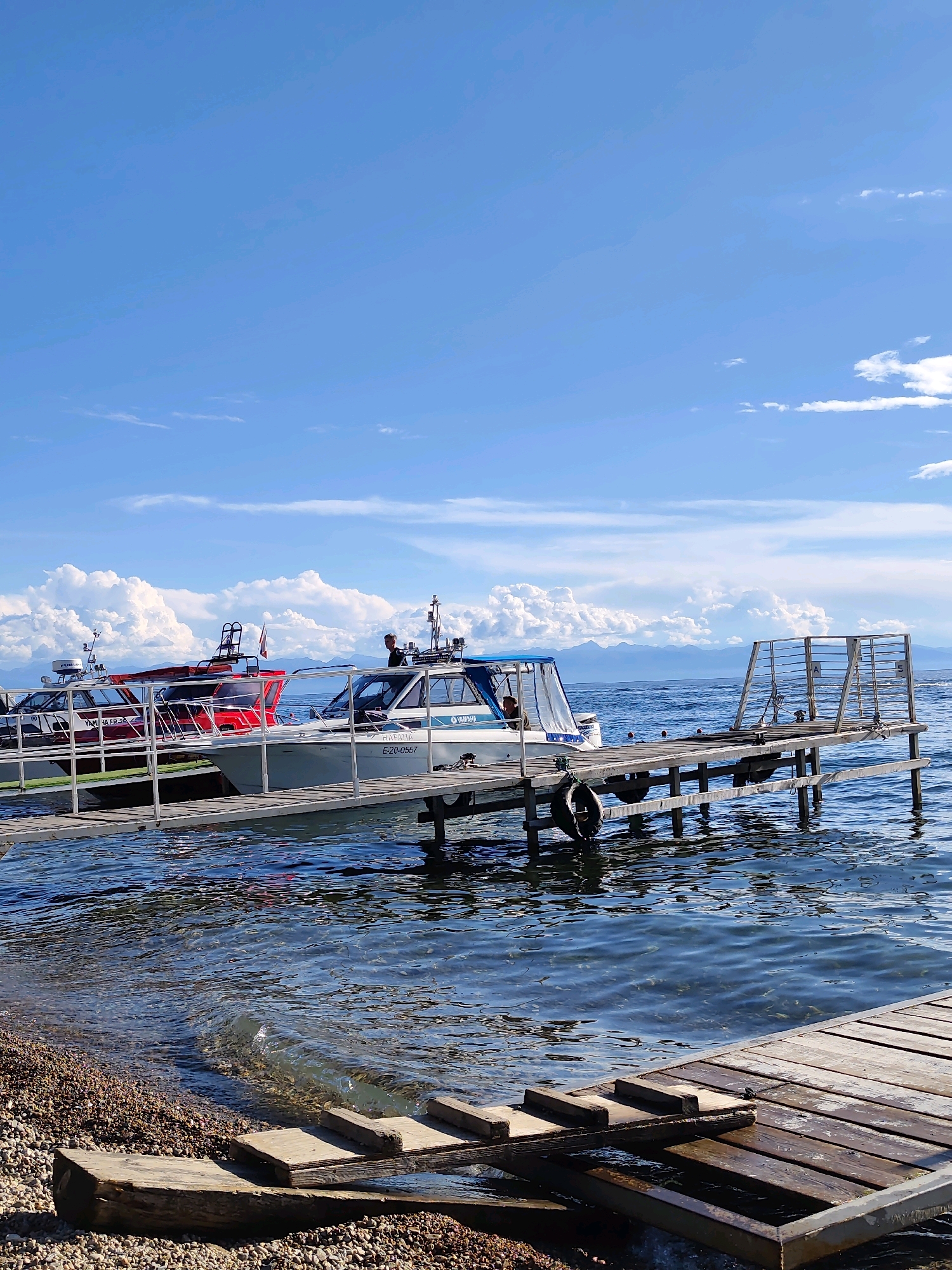 In Listvyanka, Russia, on a clear day with a bright blue sky dotted with fluffy white clouds, several boats are moored to a wooden pier on a calm body of water. Two passenger boats, one red and white, and another white with a blue canopy, are docked side-by-side. The white boat has "YAMAHA" branding and a license plate E-20-0557. A man is visible standing near the stern of the white boat, appearing to be operating it or conversing with someone. The pier extends into the water, with a metal railing and seating area at its end. The shoreline in the foreground is covered with small pebbles. In the distance, a mountain range is visible against the horizon.