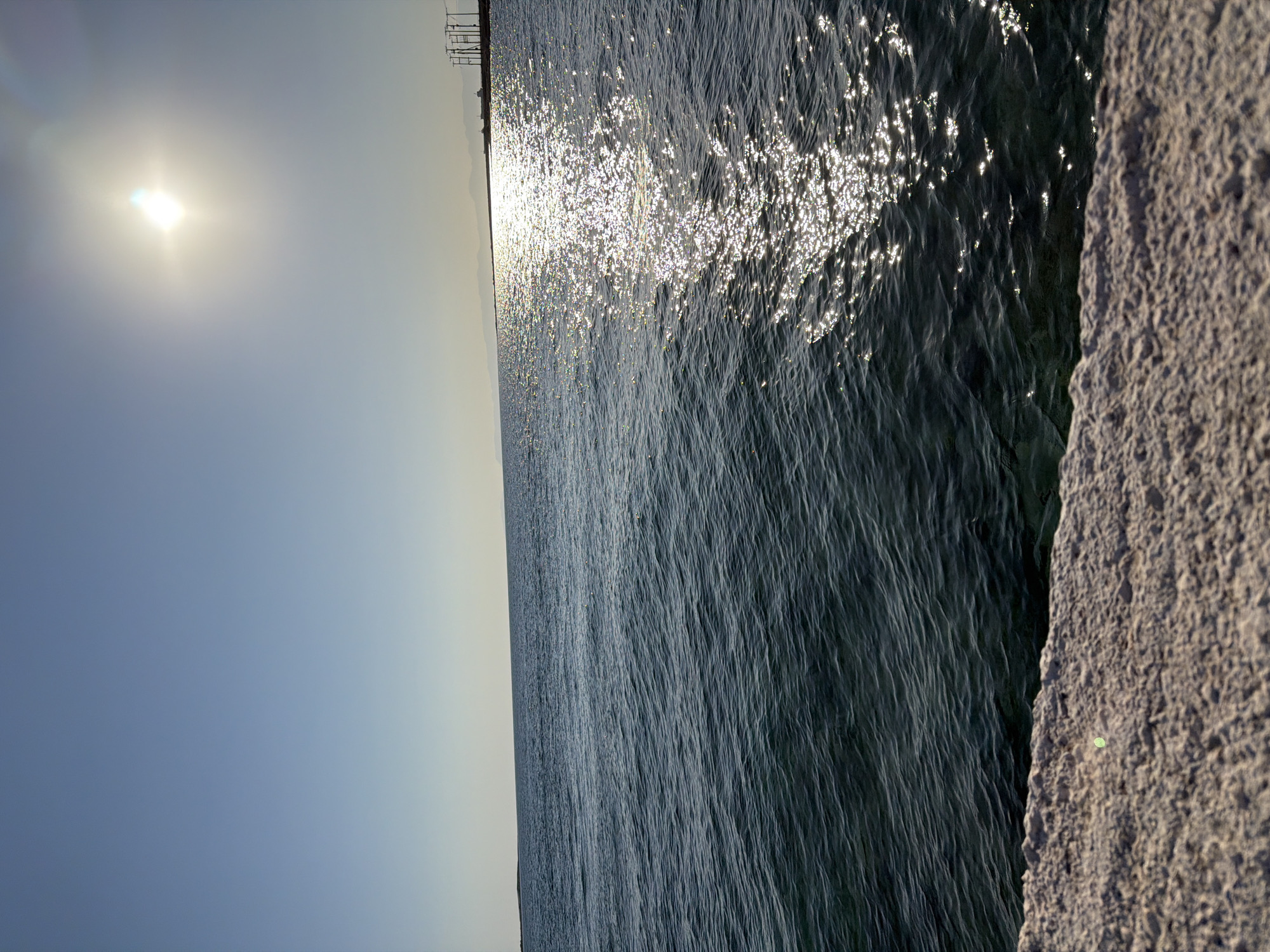The image depicts a serene seascape with a bright sun illuminating the sky and reflecting on the water. In the foreground, a textured stone embankment lines the bottom of the frame, with the rippling, dark blue ocean stretching towards the horizon. The sun, a radiant orb, is positioned in the upper right quadrant, casting a shimmering path of light across the water. In the distance, the faint outline of land and man-made structures, possibly industrial or port-related, can be seen along the coastline. The sky is a clear, gradient blue, transitioning from a lighter hue near the horizon to a deeper shade above. The scene evokes a sense of tranquility and natural beauty, characteristic of coastal areas such as Vigklakia, Greece.
