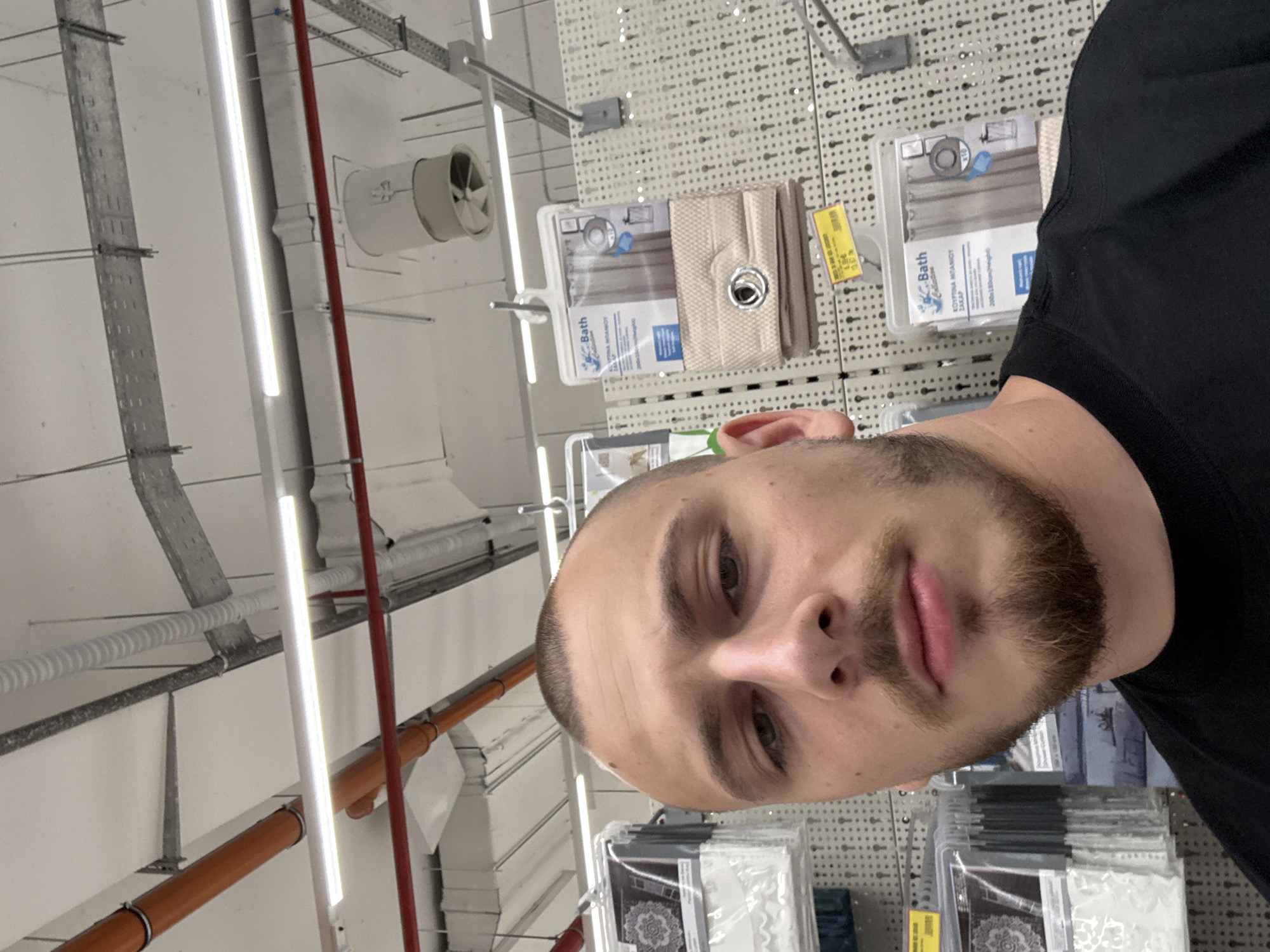 The image captures a young adult male taking a selfie or being photographed indoors, likely within a retail store in Stara Zagora, Bulgaria. The man, with a short beard and close-cropped hair, wears a black t-shirt and looks directly at the camera with a neutral expression.
The setting appears to be a commercial building with an industrial-style ceiling. Visible ceiling infrastructure includes white walls, silver cable trays, various pipes (white, red, and orange), bright fluorescent strip lighting, and a circular ventilation fan.
Behind and to the right of the man, a white pegboard display holds several packaged retail items. These items are likely bath accessories or shower curtains, with one package clearly displaying "Bath Collection" and Greek text "ΕΚΡΥΠΝΑ ΜΠΑΝΙΟΥ" (meaning "Shower Curtain") along with dimensions "200x180cm". Another package has "MADE IN CHINA" printed on it. A yellow price tag on the shelf indicates a price of "5.99 €". The image is rotated 90 degrees clockwise.