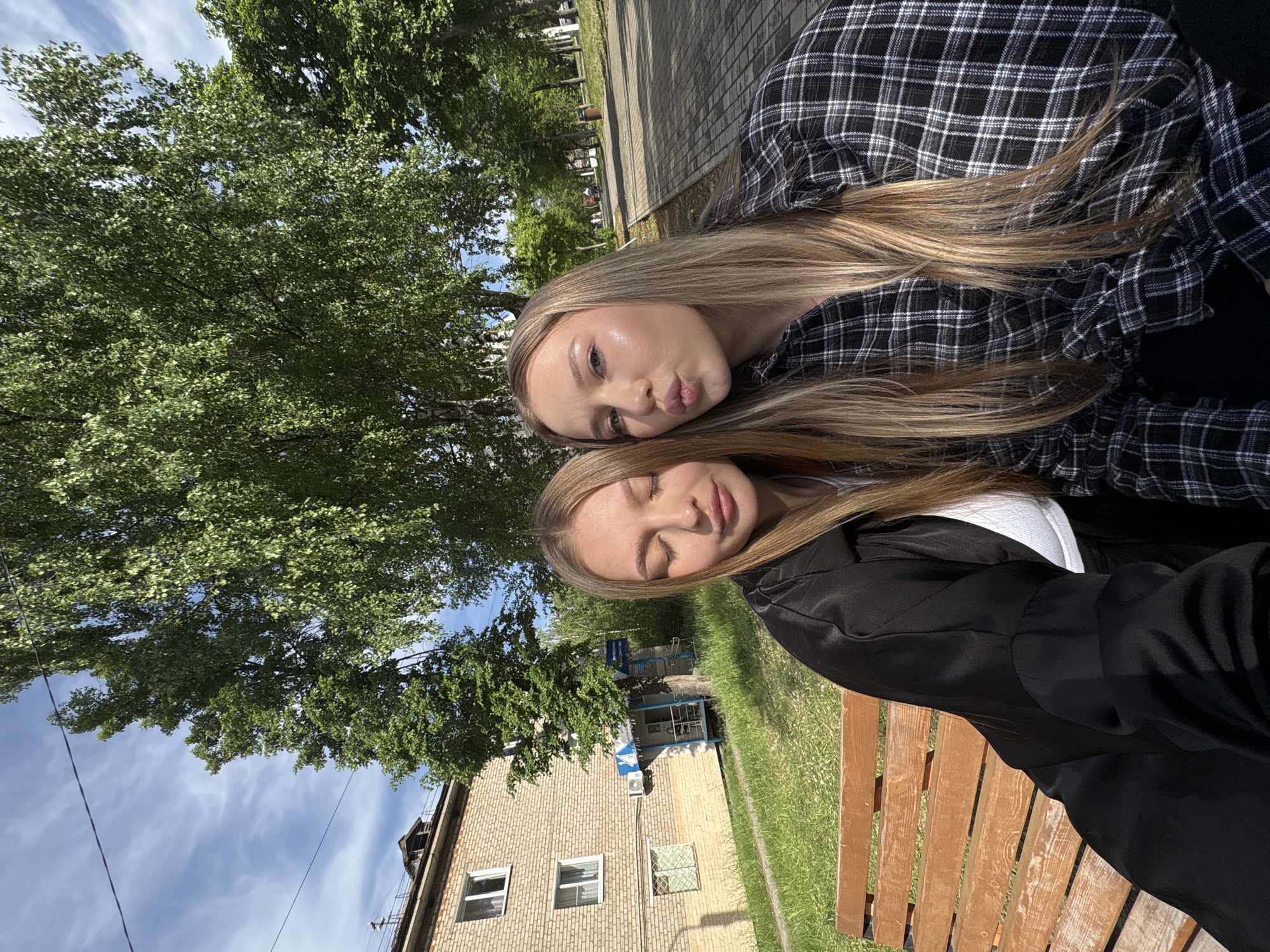 Two young women are sitting on a wooden bench outdoors. The woman on top is wearing a black and white plaid shirt and making a kissing face. The woman below her is wearing a white shirt with a black blazer. They both have long blonde hair. Behind them is a brick building and a large green tree. The sky is blue with some clouds. The location is likely Chaikovsky, Russia, due to the architectural style of the buildings and foliage.