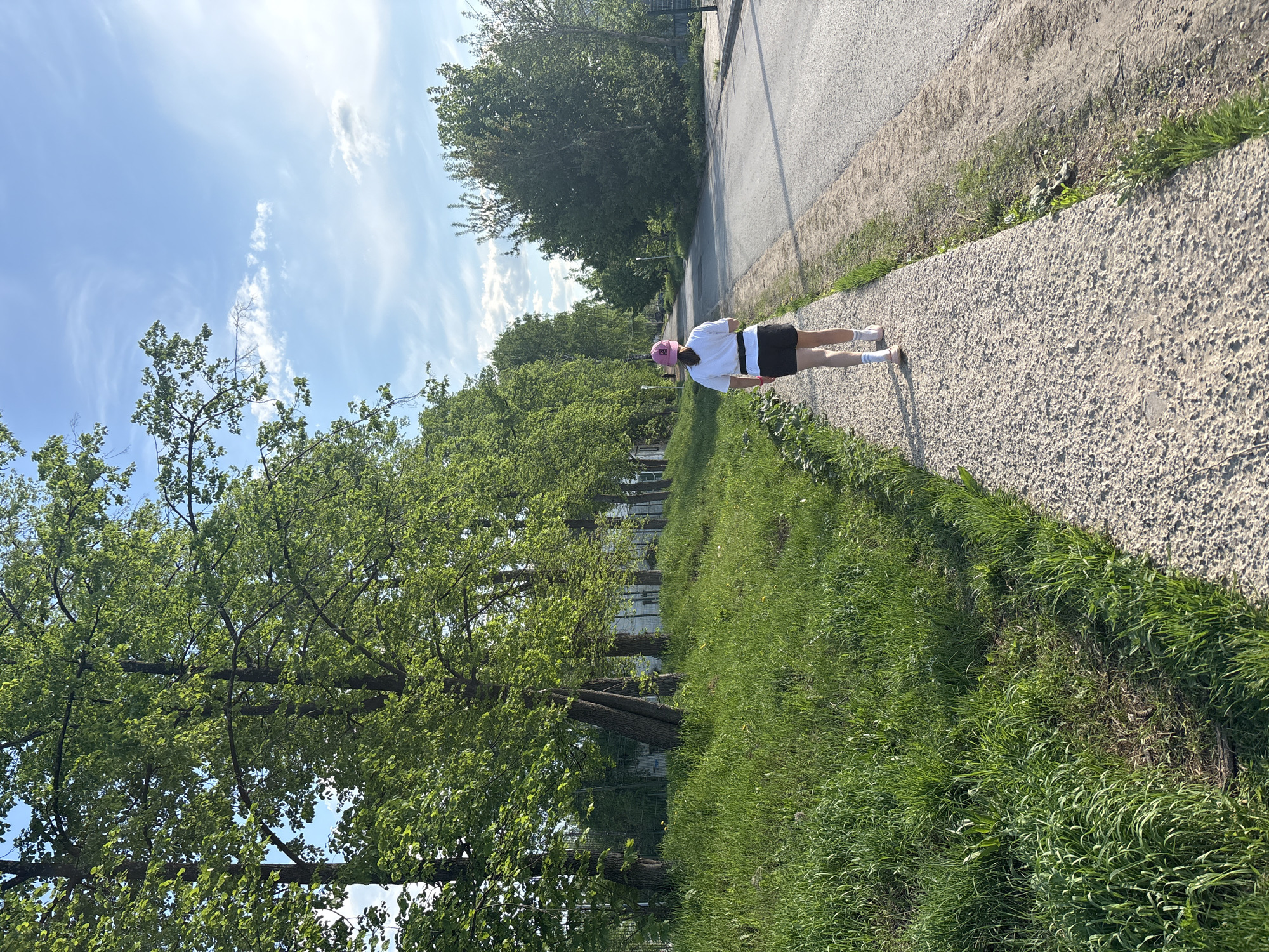 A person in a white t-shirt, black shorts, and a pink hat is walking on a sidewalk in Chaikovsky, Russia. The sidewalk is made of light gray gravel and runs alongside a road. To the left of the sidewalk is a grassy area with trees, and in the background, there are buildings. The sky is blue with scattered white clouds.